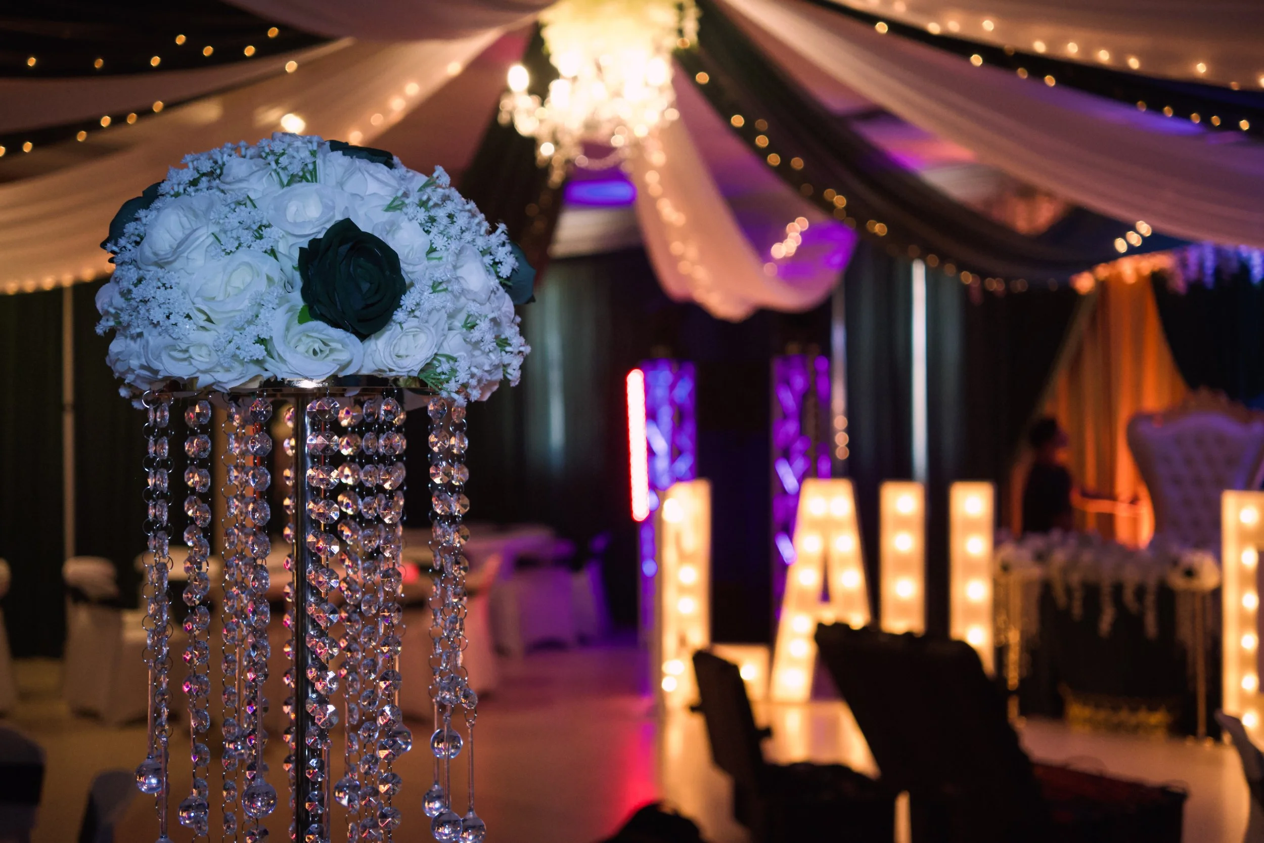 A floral centerpiece with white and black roses hanging crystal decorations, set against a backdrop of a decorated event space with draped fabric, string lights, and lit candles.