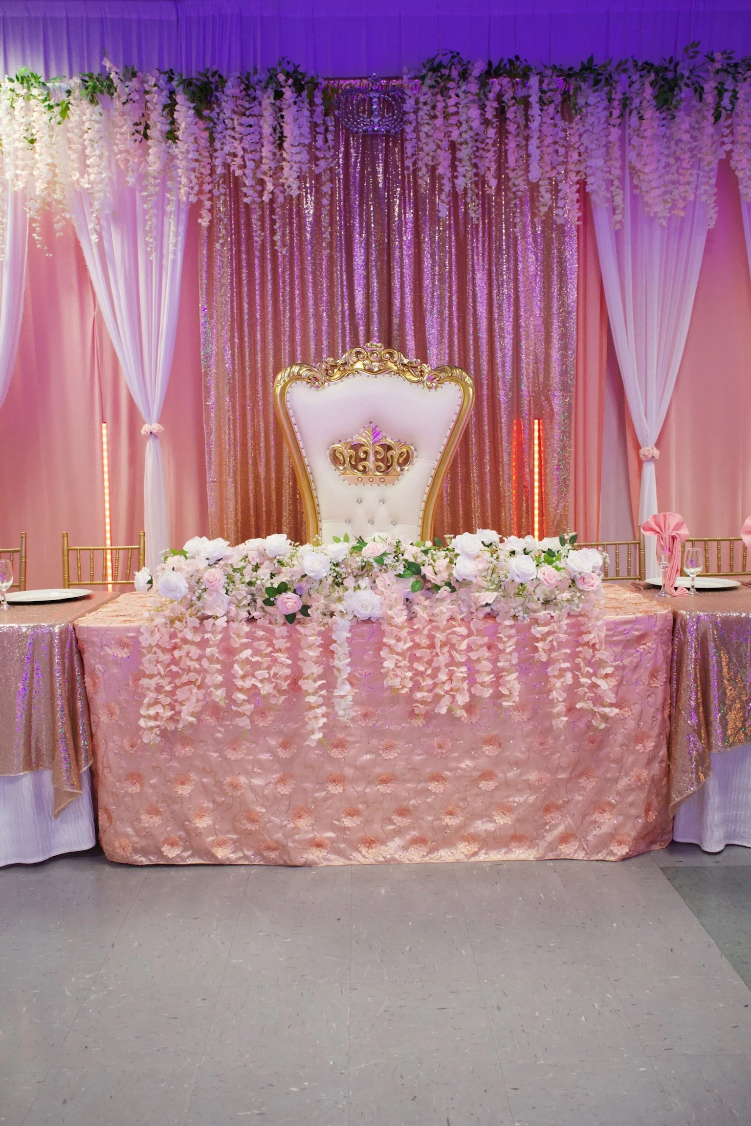 A decorated wedding or special event table with a floral arrangement, a pink tablecloth, and a backdrop of pink and purple hanging beads, white curtains, and floral garlands.