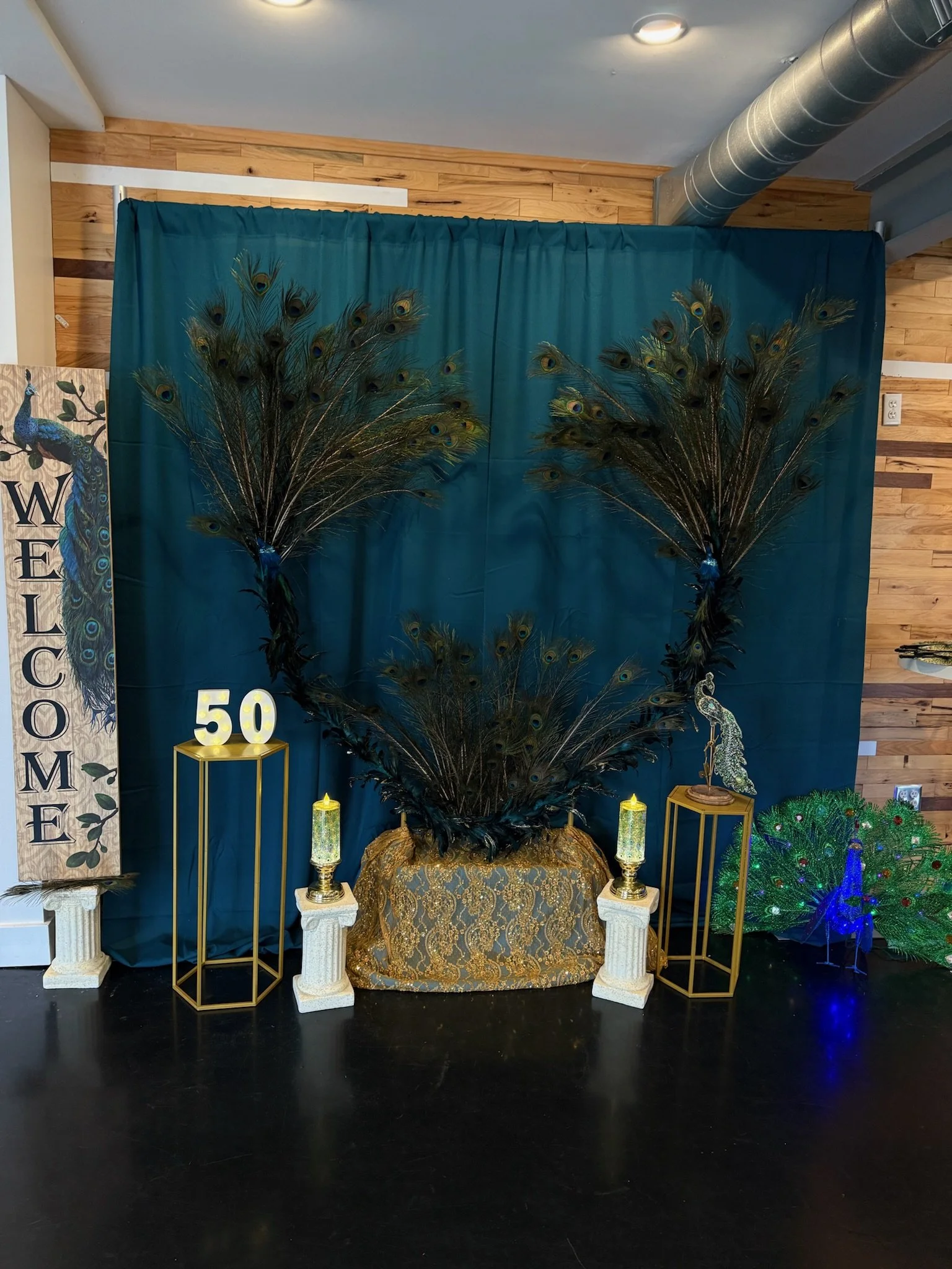 Decorative backdrop feature with peacock feathers, gold and white pillars, teal curtains, and illuminated number 50, set up for celebration or event.