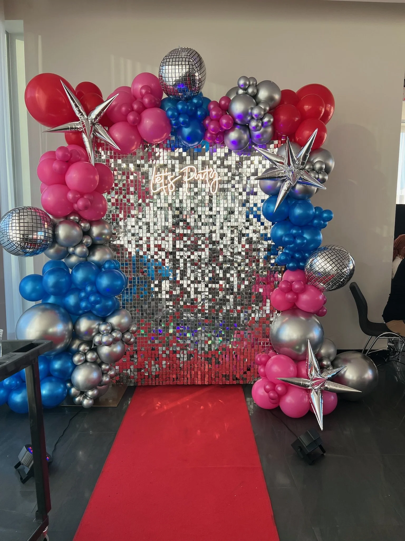 Balloon arch and backdrop for a celebration event with pink, blue, silver, and star-shaped balloons, and a sign that says 'Let's Party' on a silver mirror mosaic background.