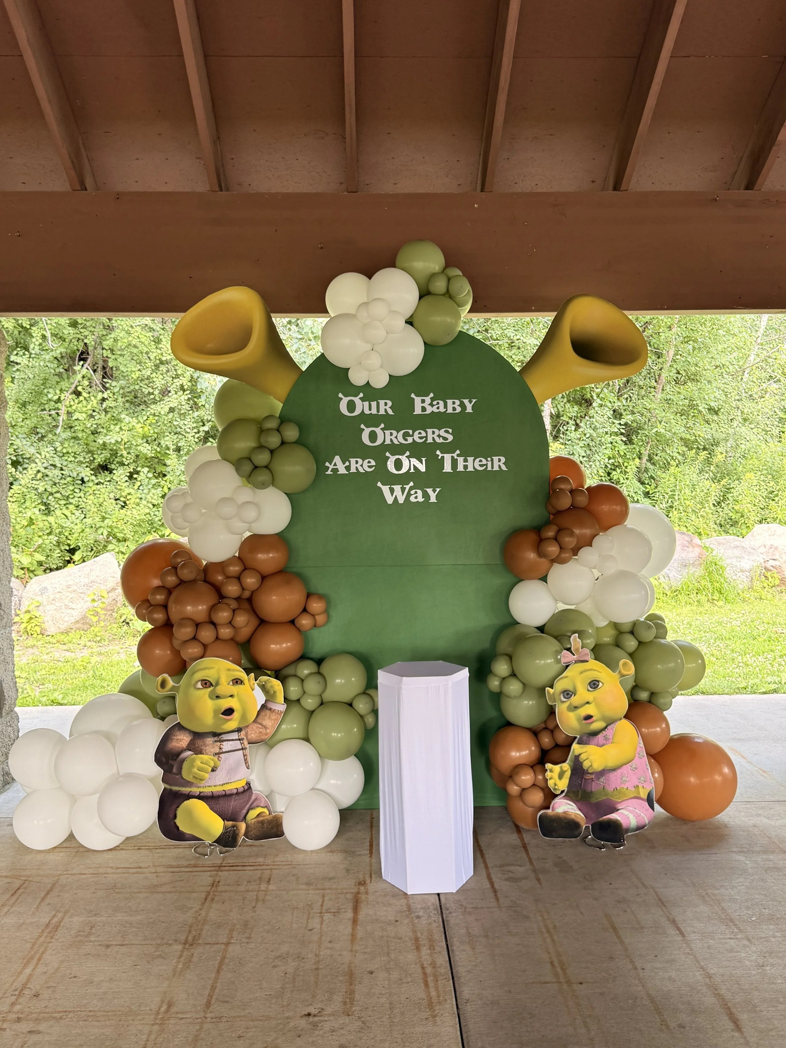 Decorative display for a baby shower with green, white, and brown balloons, Shrek and Fiona character cutouts, and a green backdrop with the message "Our Baby Örgörs Are On Their Way."