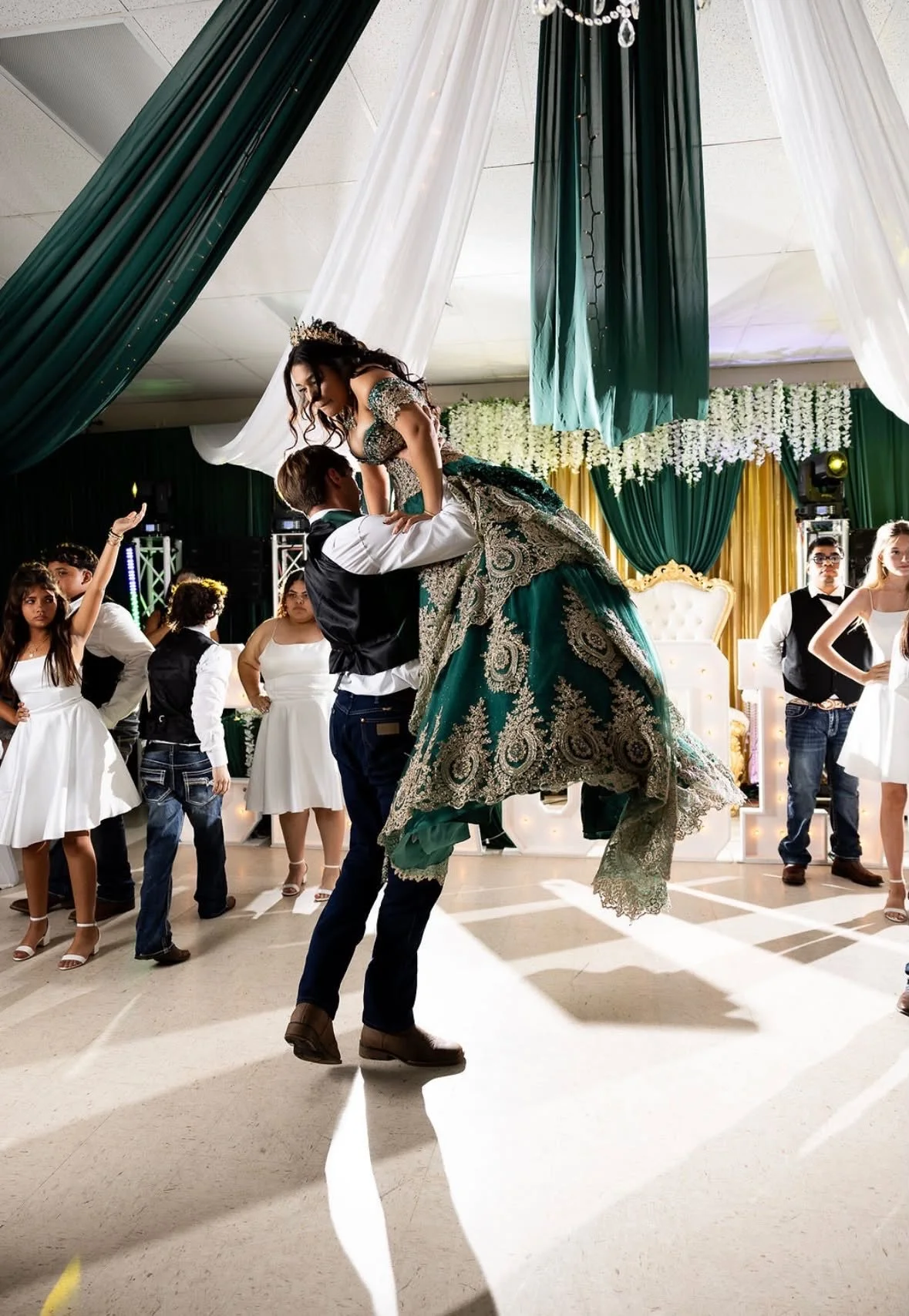 A young woman in a green and gold traditional dress is lifted by a young man during a celebration or dance. She appears to be wearing a crown. Several other people, mostly children, are watching or participating, with some dressed in white dresses an