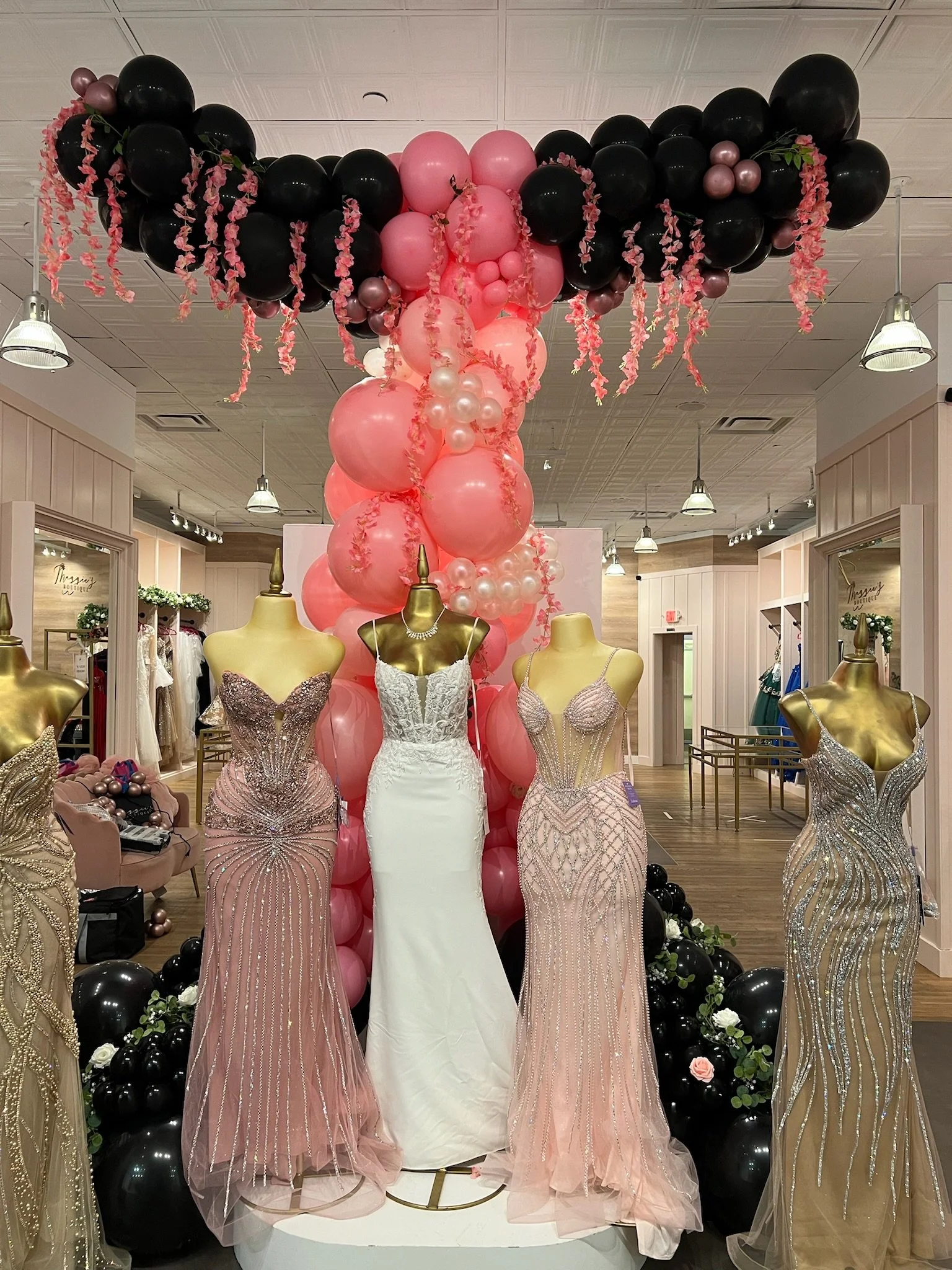 Display of wedding dresses on mannequins, surrounded by black and pink balloons, with pink floral decorations hanging from the balloons.