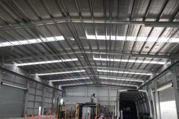 Inside a large industrial or warehouse building with a high arched metal ceiling and walls, featuring garage-style doors and parked vehicles, including a white van and other trucks.