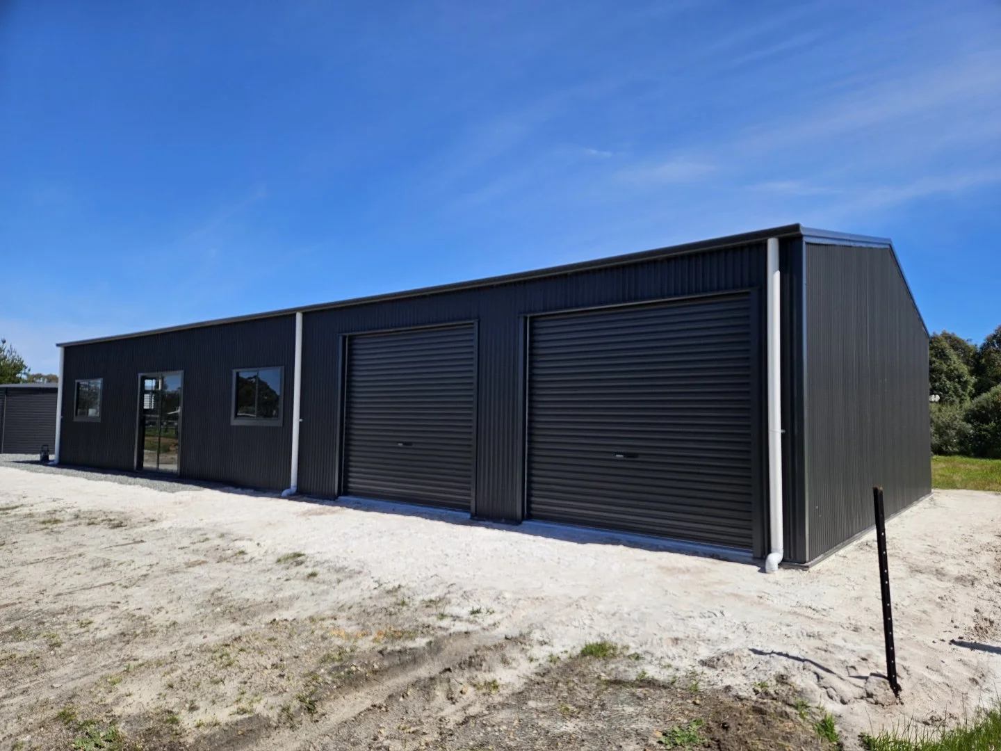 Black metal industrial storage building with two roll-up doors and windows, on a gravel lot under a clear blue sky.