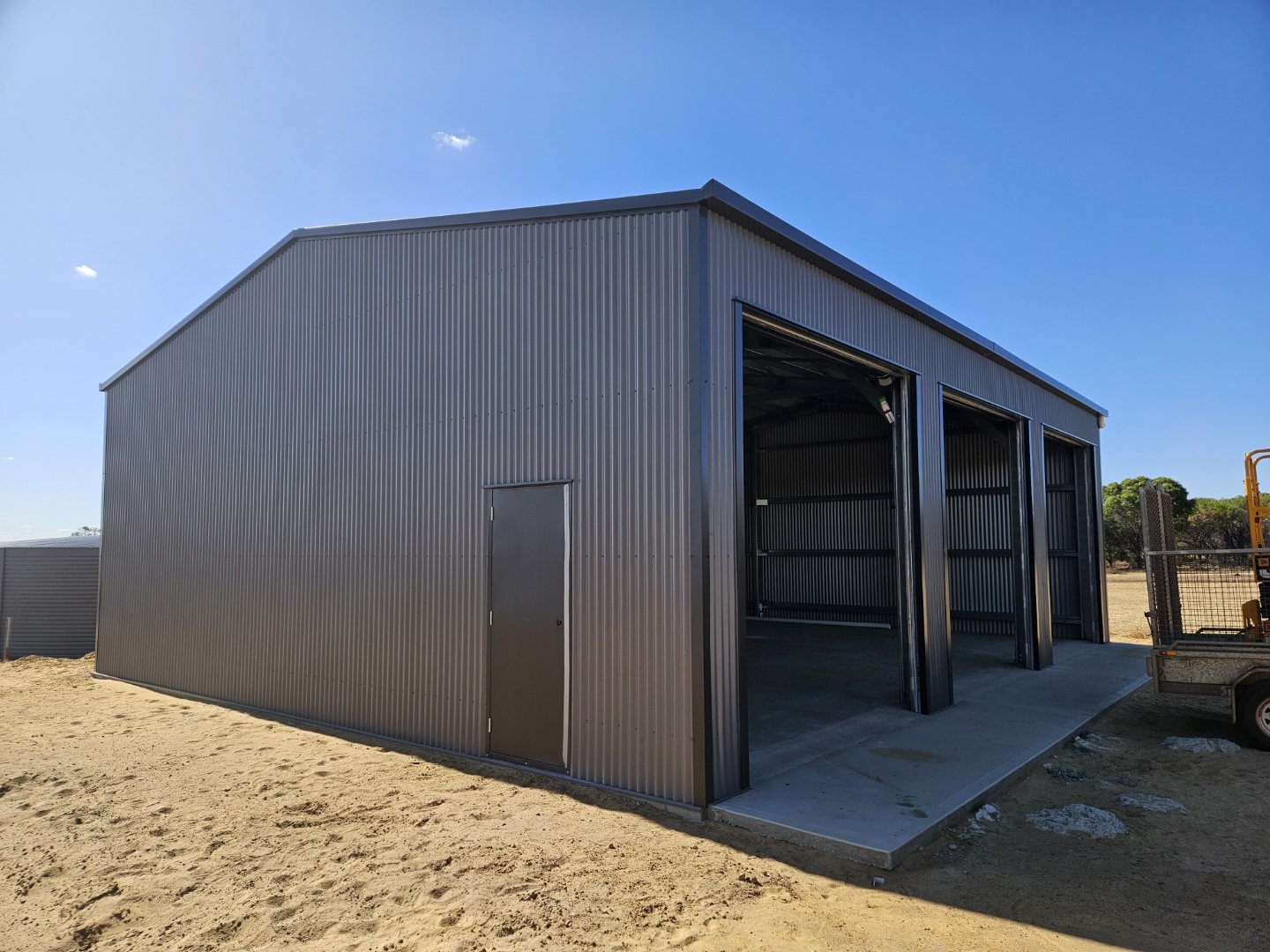 A metal storage shed with open bays and a closed door, situated on a sandy ground under a clear blue sky.