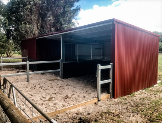 A small outdoor animal shelter with a sandy area, metal fencing, and a red corrugated metal roof and walls.