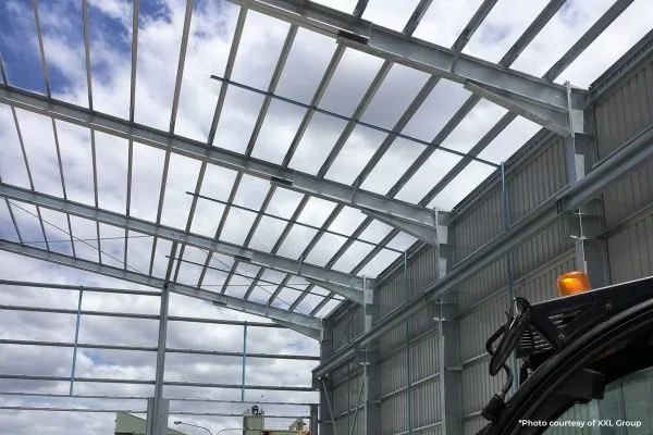 A large industrial or agricultural building with a metal framework and a partially completed glass roof, showing blue sky and clouds outside.