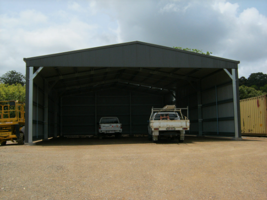 A metal storage shed with two white pickup trucks parked inside, outdoors on a dirt lot with trees in the background under cloudy skies.