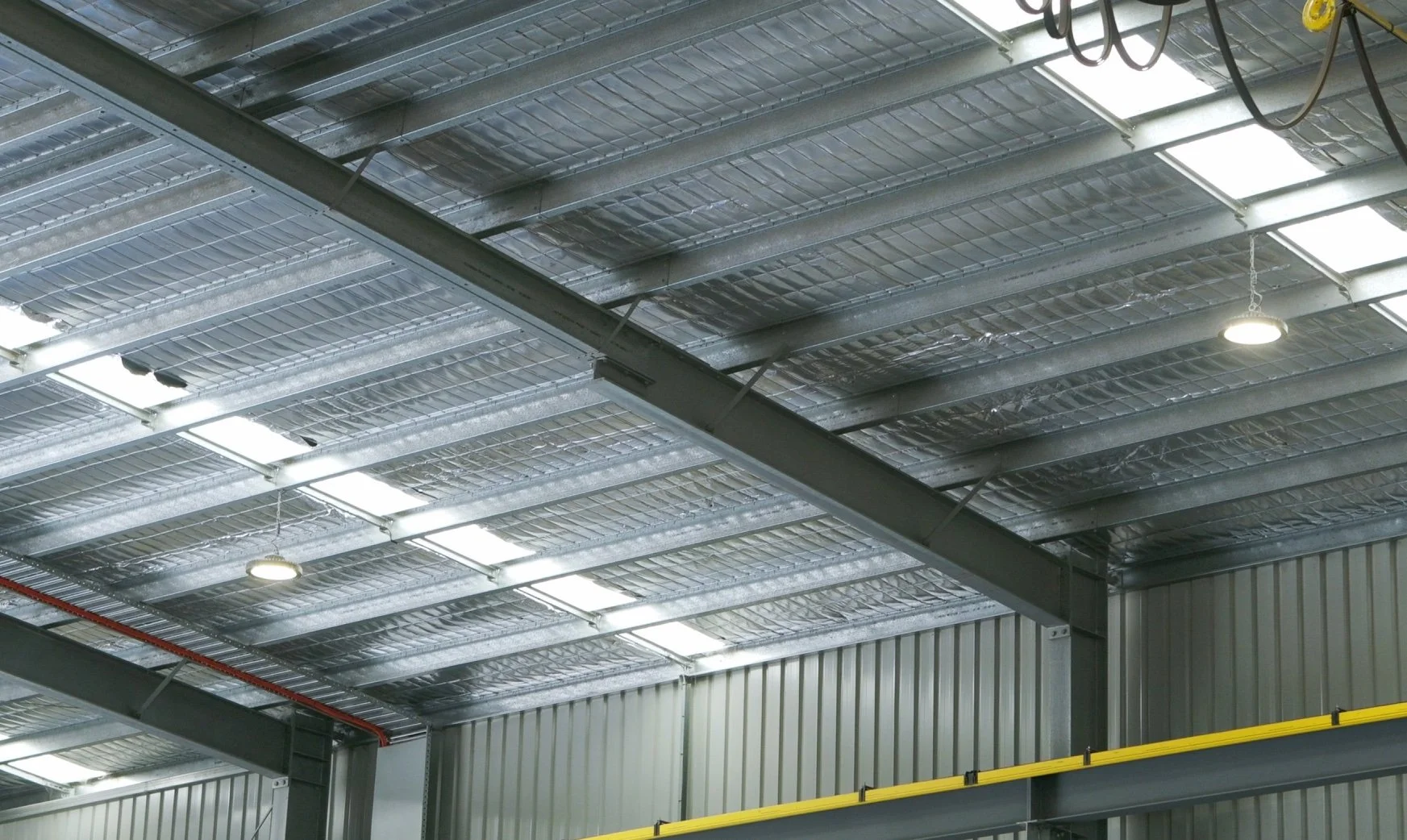 Interior view of an industrial warehouse ceiling with metal beams, insulation, skylights, and overhead lighting.