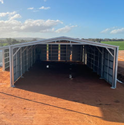 Steel-framed building under construction on a dirt lot, with a clear blue sky and distant fields in the background.