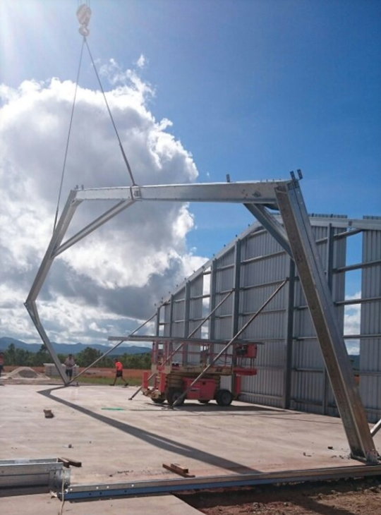 Construction site with a large metal frame structure being assembled next to a tall curved metal wall. A scissor lift is positioned underneath the structure, and a crane is lifting or supporting part of the framework. There are a few workers in the background under a partly cloudy sky.