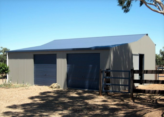 Metal shed with two closed garage doors and a side door, surrounded by dirt and fencing, under clear blue sky.