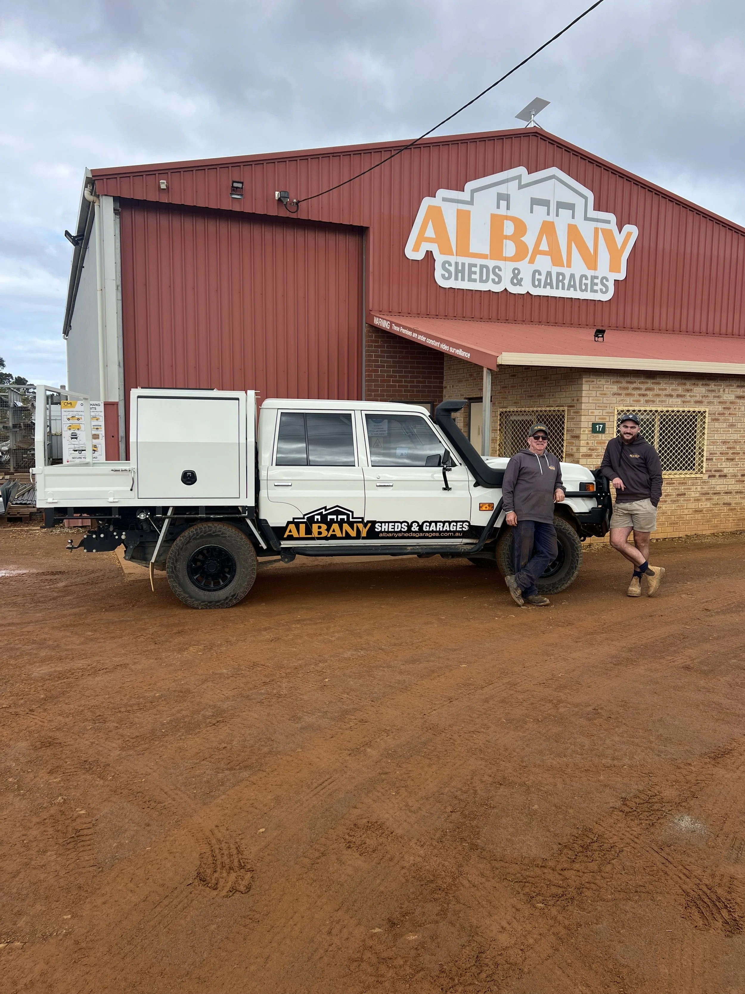 A red building with a sign that reads 'ALBANY SHEDS & GARAGES', with two people standing in front of a white utility truck parked on a dirt ground.