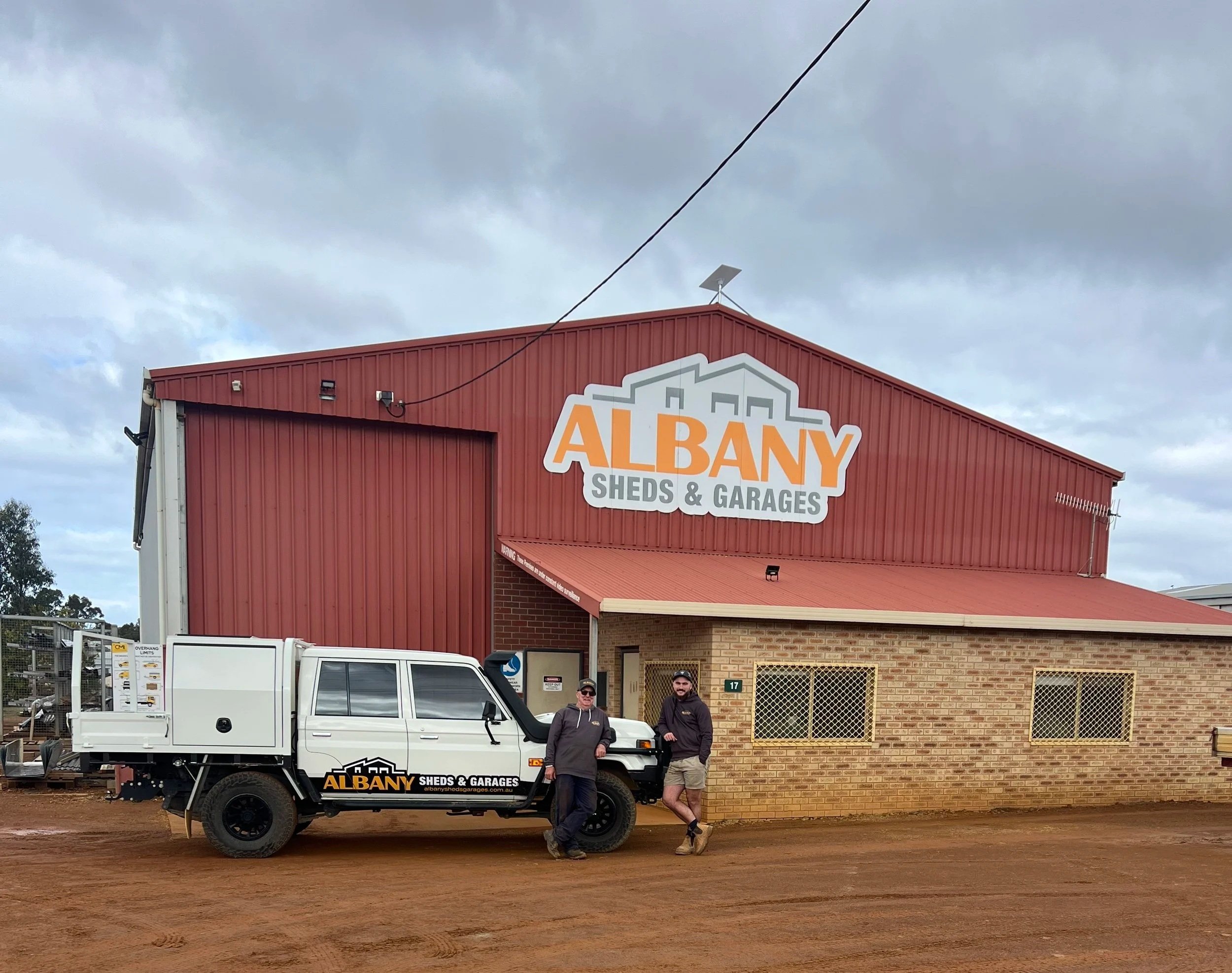 Two men standing in front of a white truck outside a building with a sign that reads 'ALBANY SHEDS & GARAGES.' The building has a red metal roof and brick walls. There are cloudy skies above.