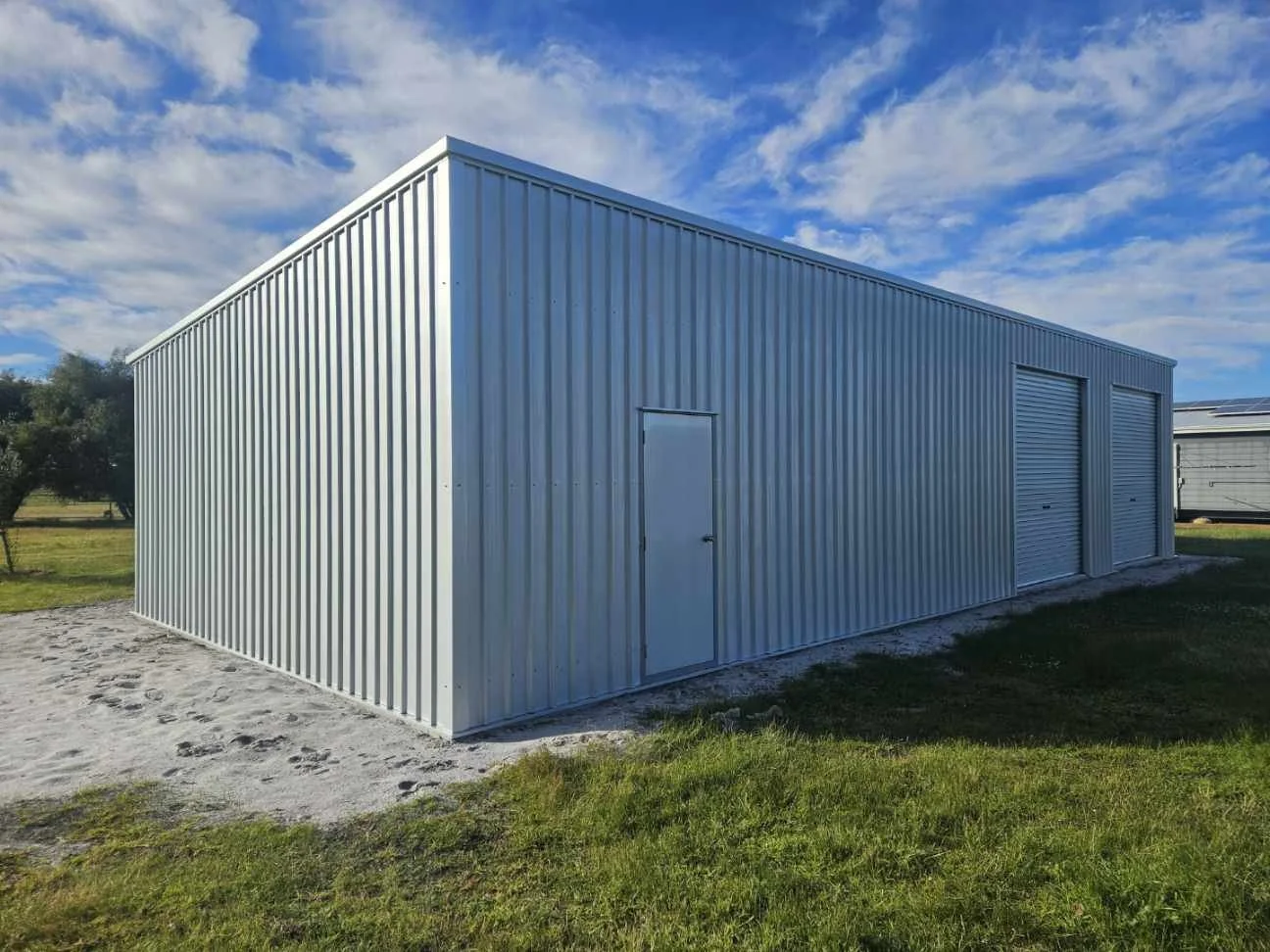 White metal storage building with a small side door and two large roll-up doors, situated on a grassy area under a partly cloudy sky.