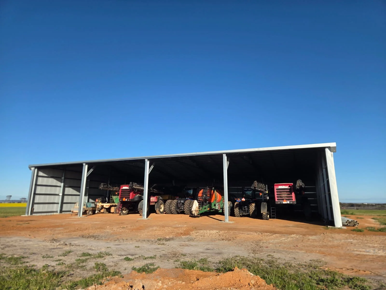 Farm equipment stored inside a large outdoor metal shed on a farm, with a clear blue sky and open fields in the background.
