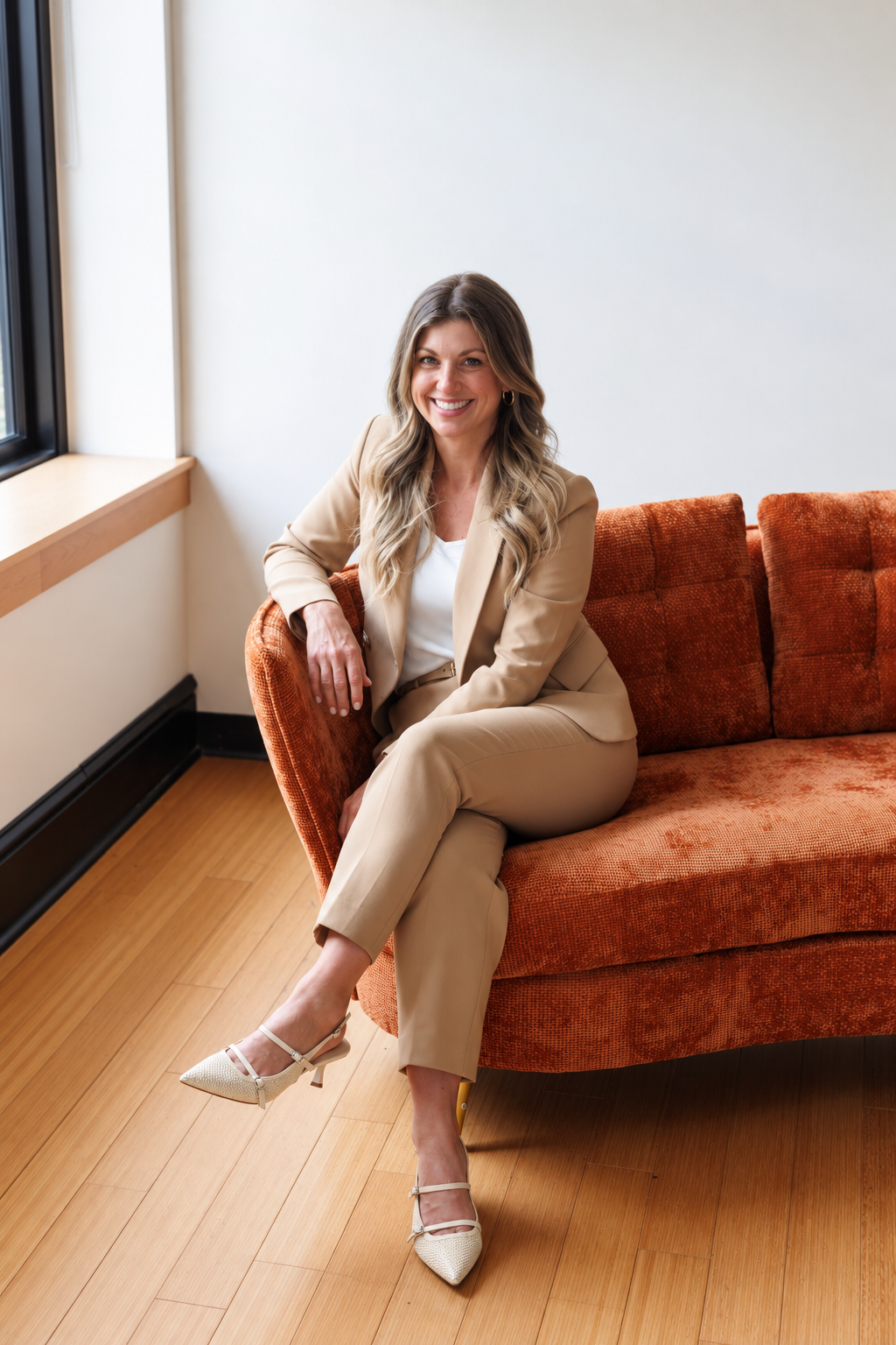 A woman with blonde hair sitting on an orange sofa by a window, smiling, wearing a beige suit and white top, with beige heels.