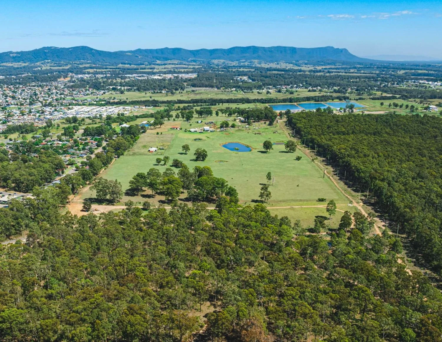 Aerial view of a Werakata Estate with green fields, small ponds, clusters of trees, and a forested area on the right, with Cessnock town centre in the distance and mountains in the background.