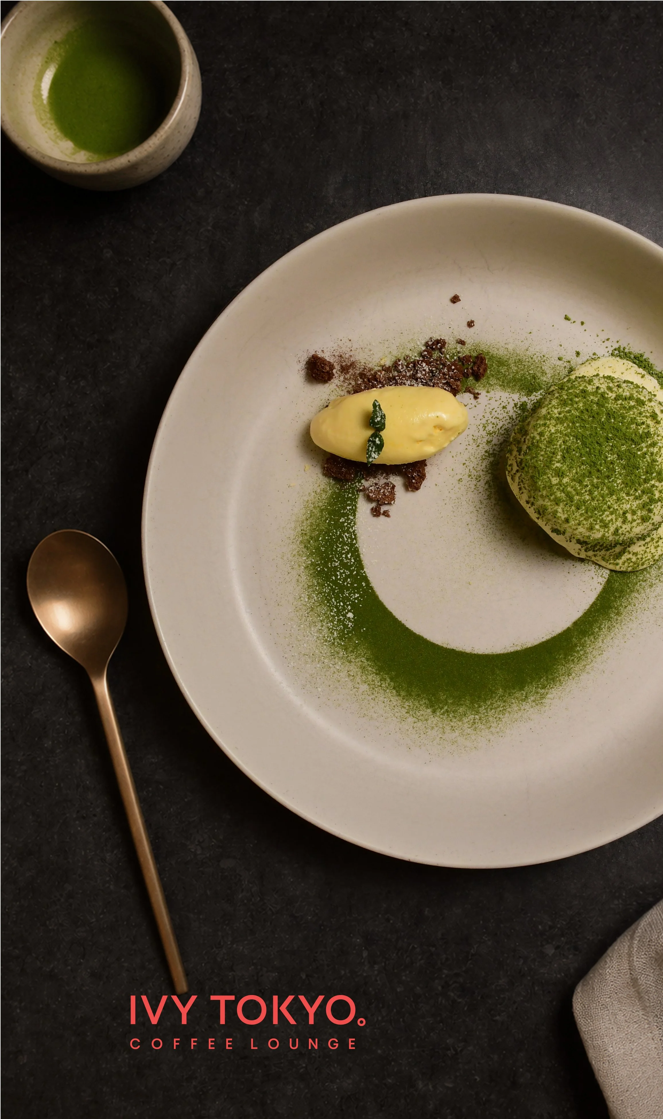 A white plate with green matcha powder, a scoop of ice cream with green topping, and small brown crumbs on a black surface. A small cup of matcha tea is in the top left corner, and a gold spoon is placed to the left of the plate. The words 'IVY TOKYO. COFFEE LOUNGE' are written in red at the bottom.
