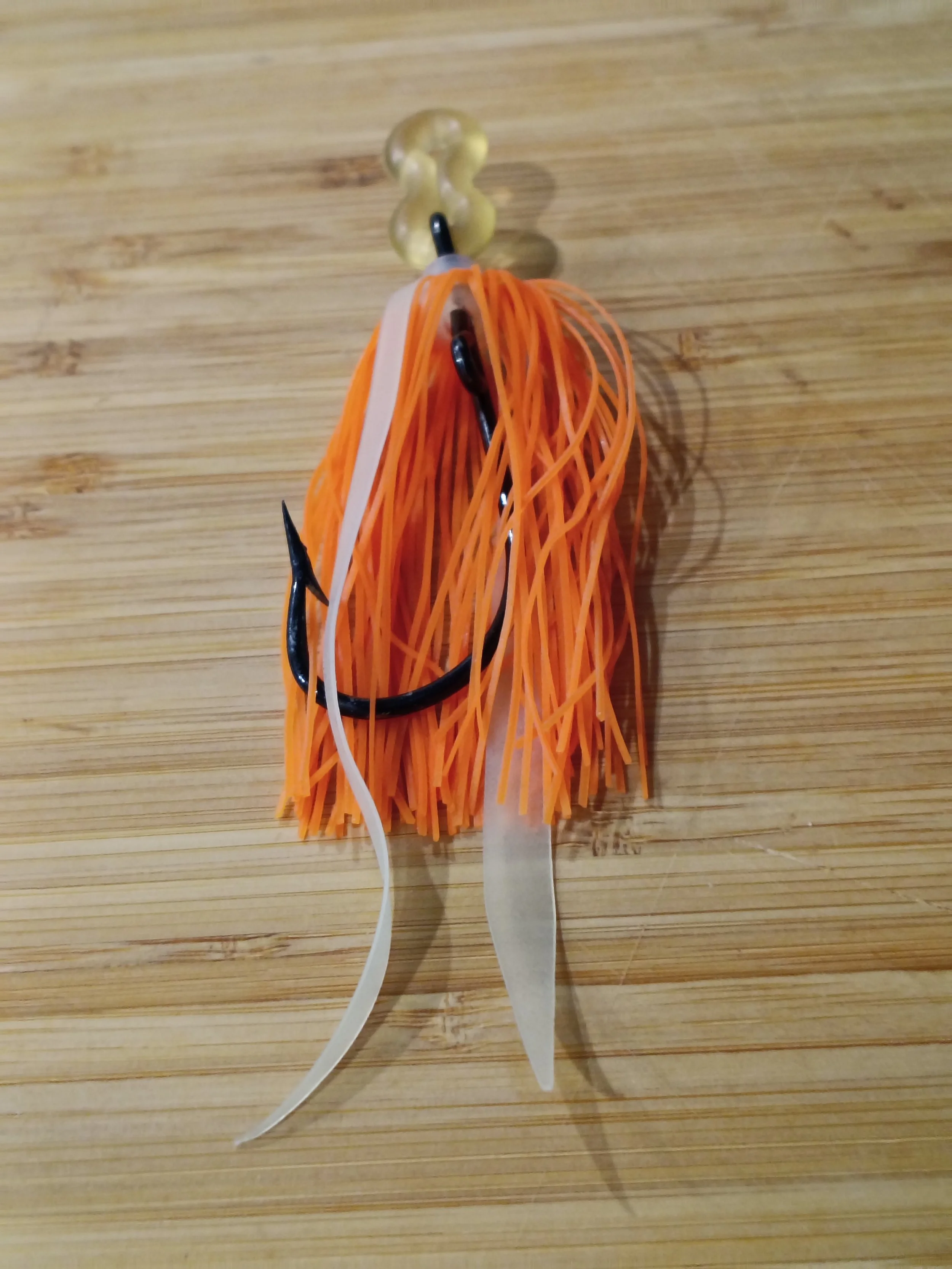 A fishing lure with orange rubber strands, black hooks, a white plastic tail, and a transparent top with yellowish beads, hanging on a wooden surface.