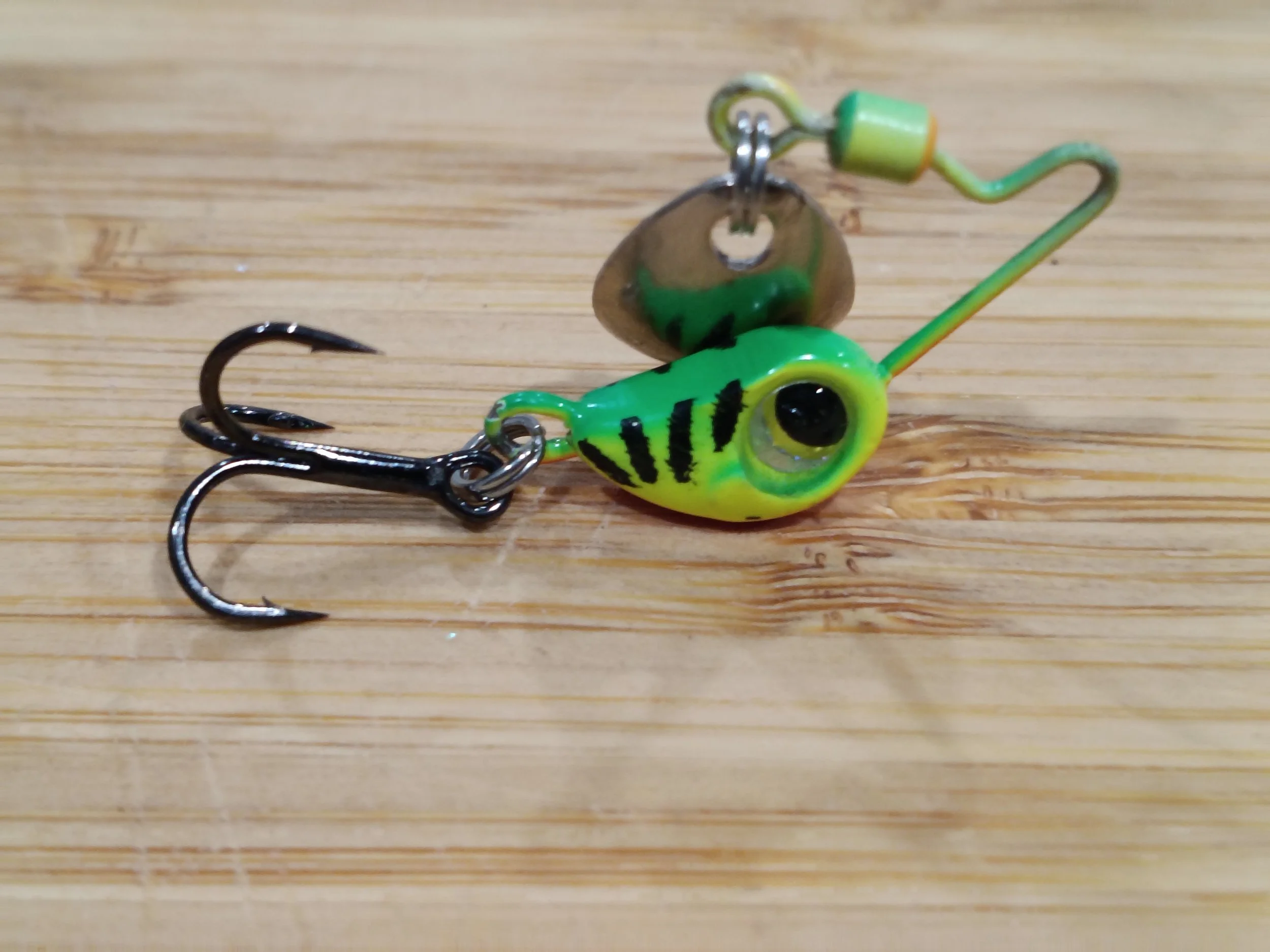 Close-up of a small fishing lure with a green and black striped fish-like design, a metal spinner blade, and a treble hook, placed on a wooden surface.