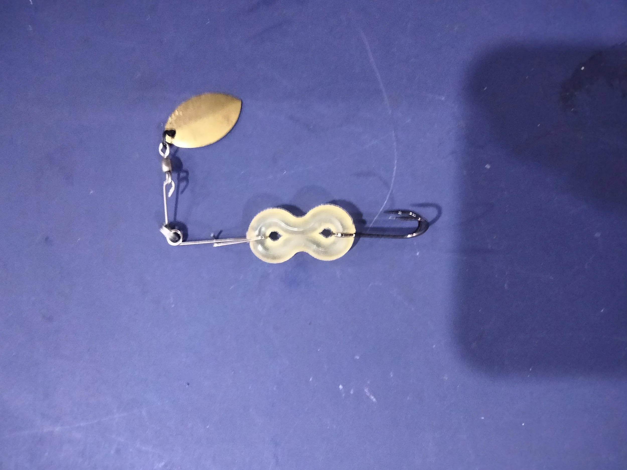 A close-up view of a fishing lure with a gold fish-shaped body, a black eye, and a clear plastic body with a metal hook attached, against a dark blue background. Incorporating fishooka figure eight hooking system. finnovations.shop