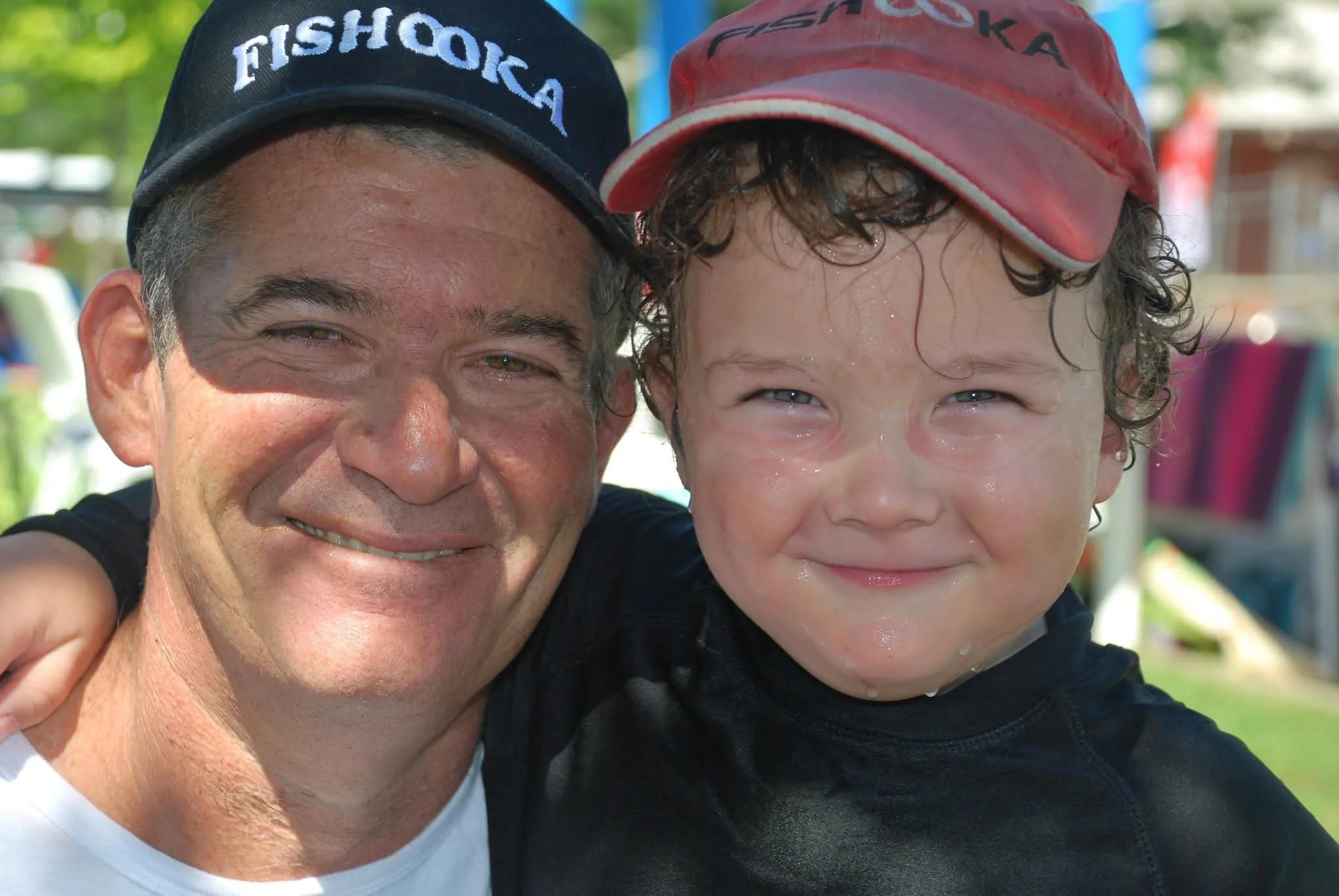 A man and a young boy are smiling and close together, with the man holding the boy. The man is wearing a black cap that says 'FISH POLKA,' and the boy is wearing a red cap. They are outdoors on a sunny day.