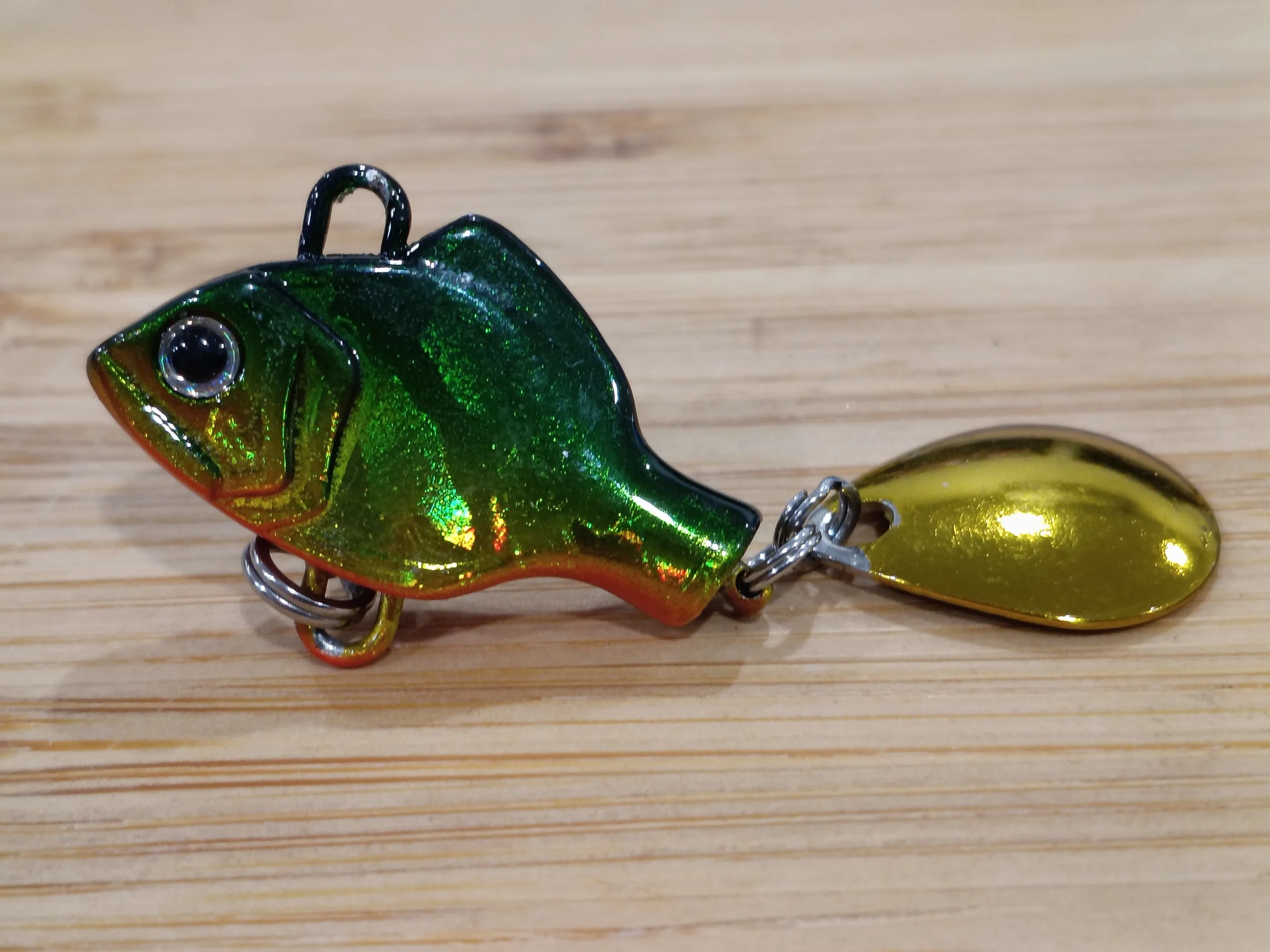 Close-up of a fishing lure shaped like a frog, with a shiny green and black body, a large eye, and a metal hook.