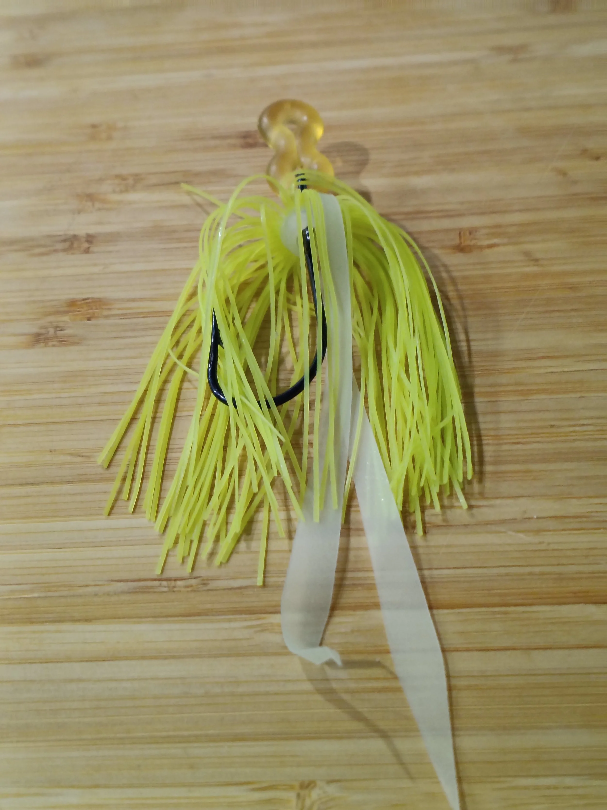 Close-up of a fishing lure with yellow strands, a white strip, and a black hook, placed on a wooden surface.