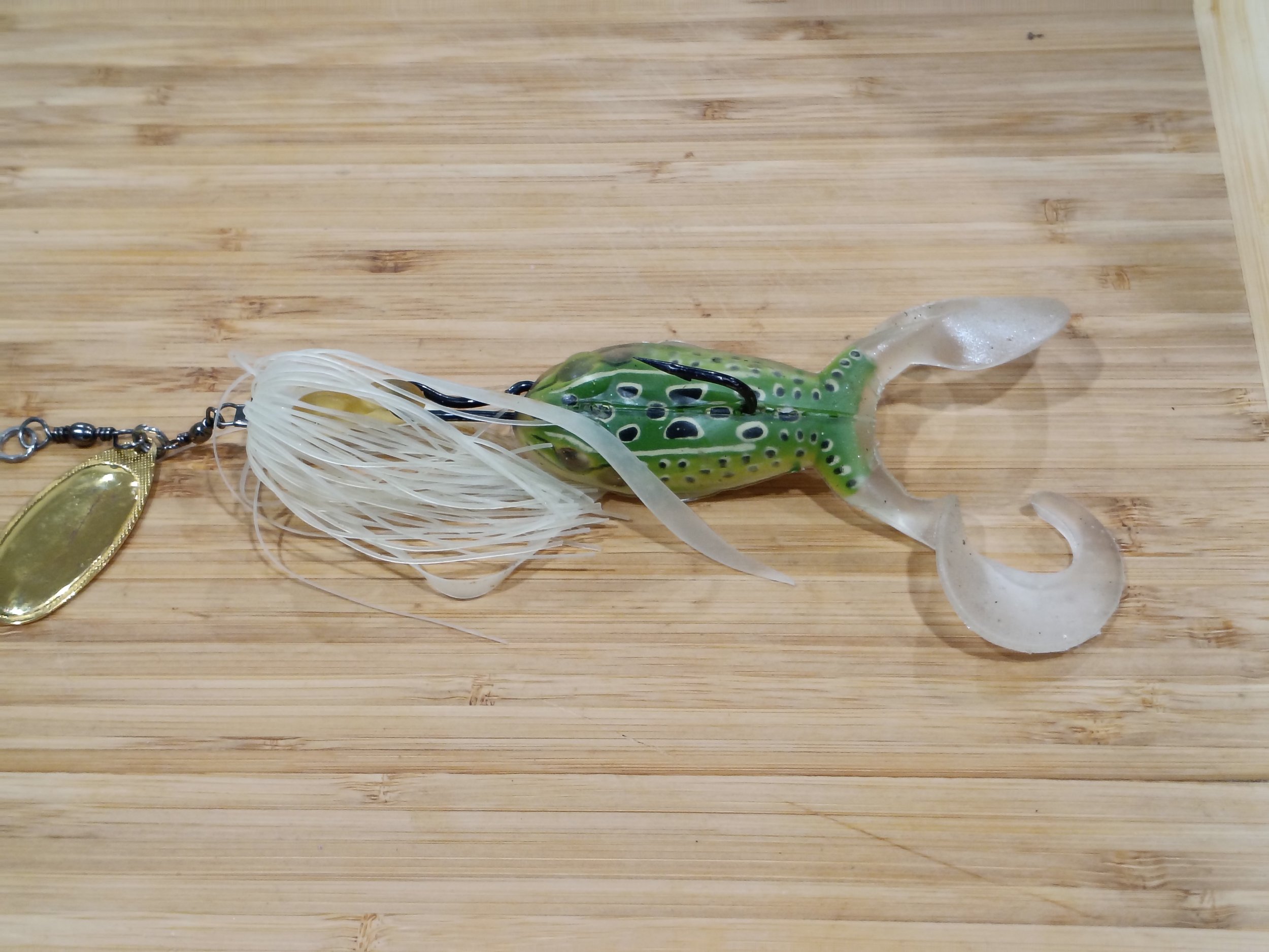 A fishing lure designed to resemble a small sea creature with a green and black spotted body, a clear tail, white hair-like appendages, and attached to a fishing line and hook.