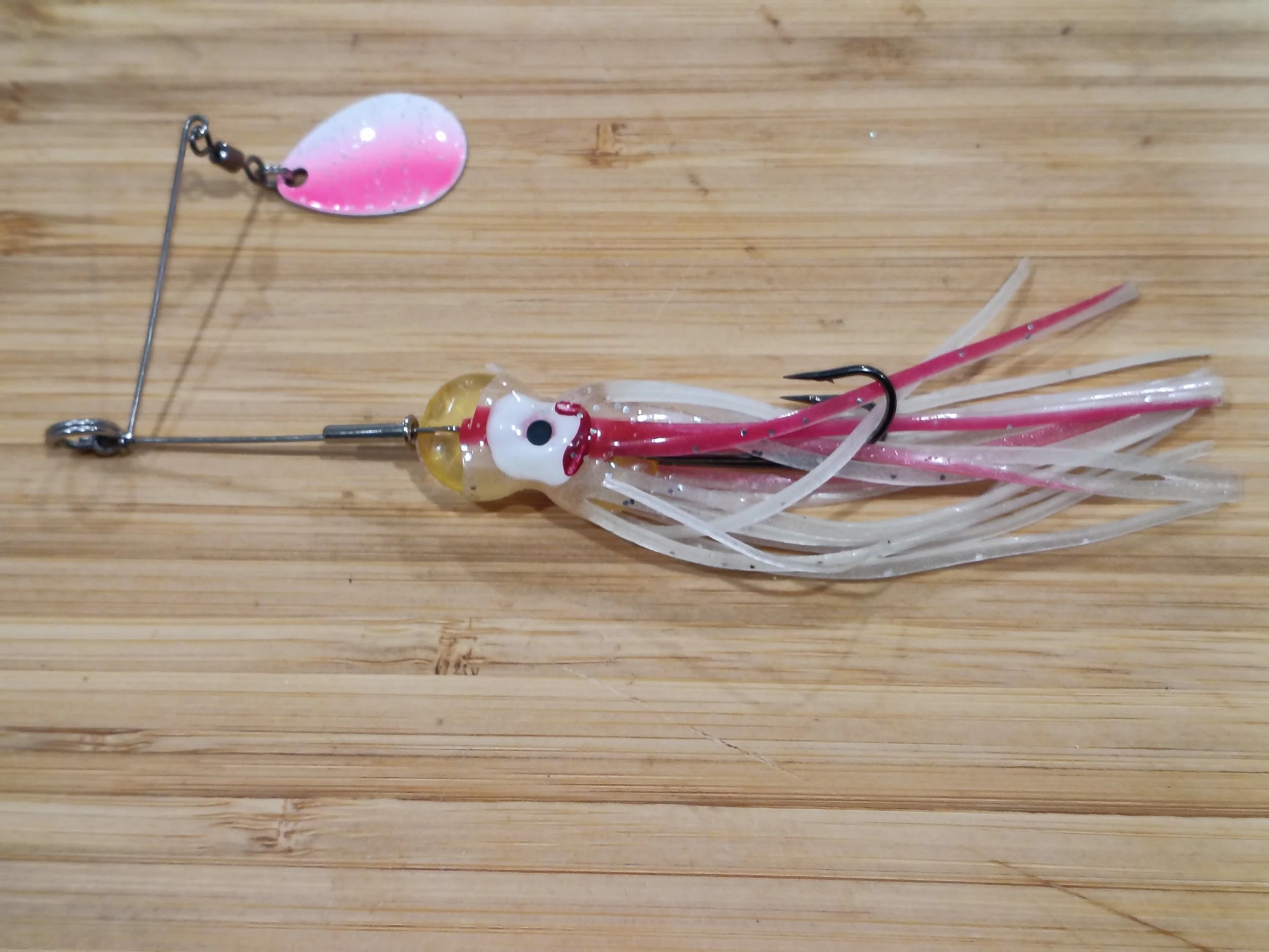 A fishing lure with a pink and white oval-shaped top and a plastic squid-shaped body with pink, white, and yellow colors, lying on a wooden surface.