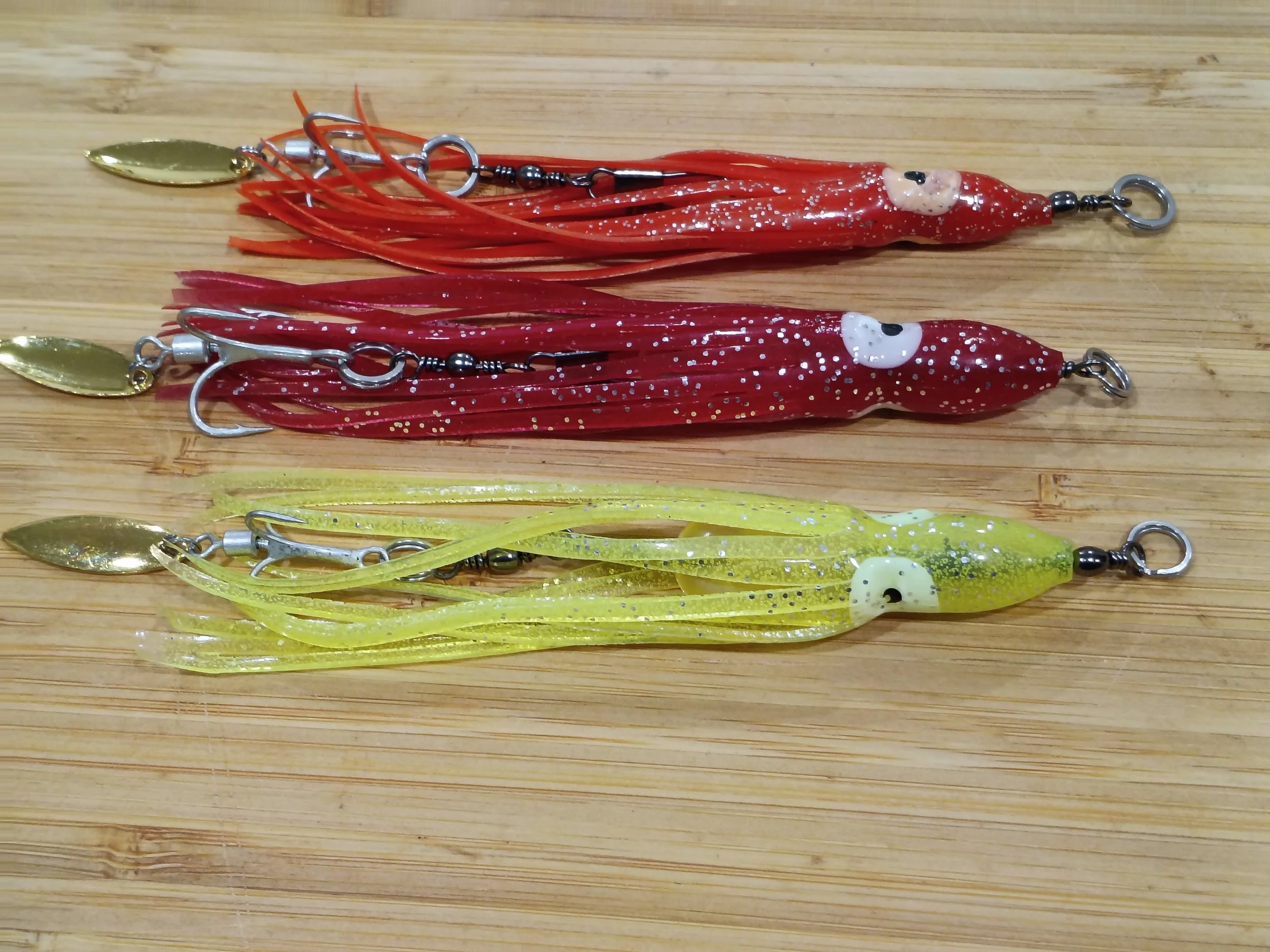 Two fishing lures shaped like squid, one red with glitter and one yellow with glitter, laid on a wooden surface.
