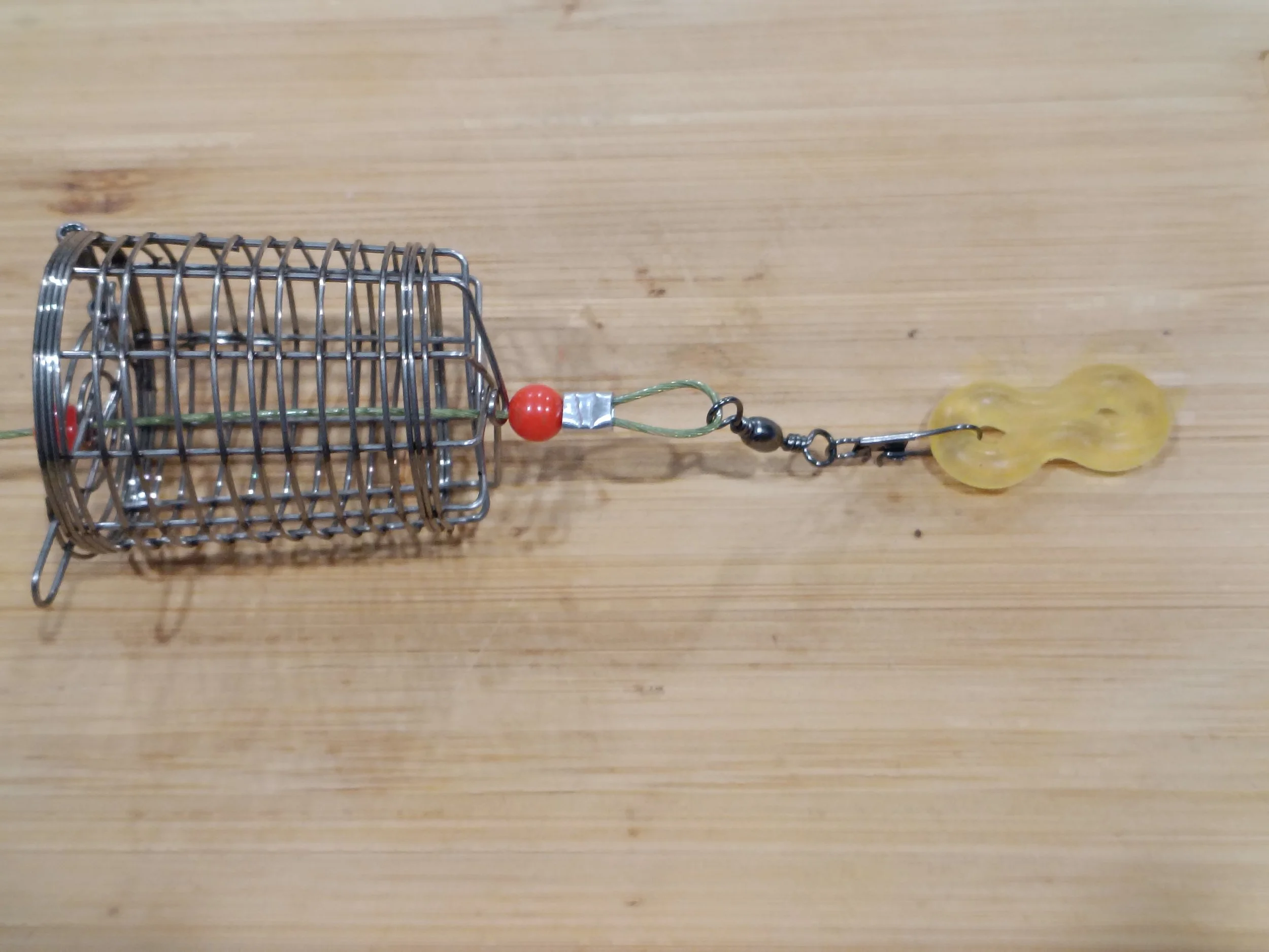 A small metal wire cage with a red bead connected to a yellow plastic lure on a wooden surface.