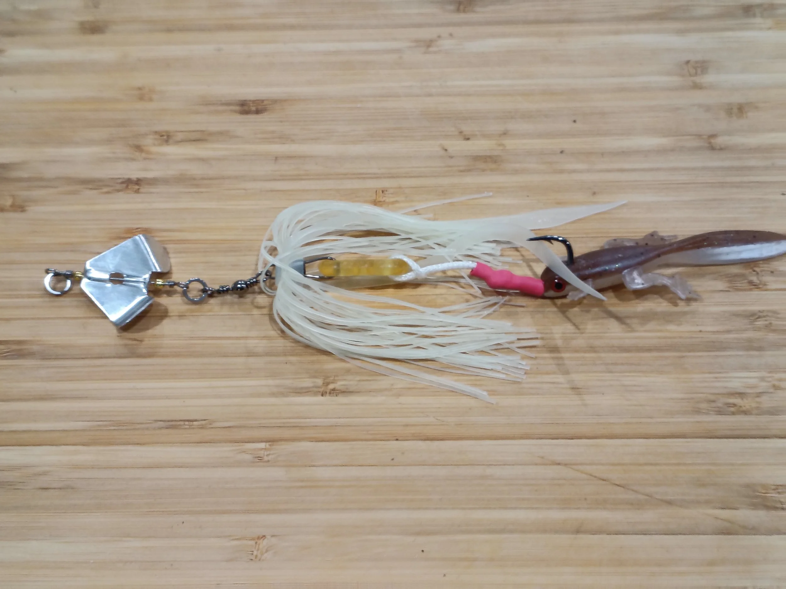Fishing lure with a fish-shaped soft plastic body, feathered skirt, and metal clip attached to a light-colored wooden surface.