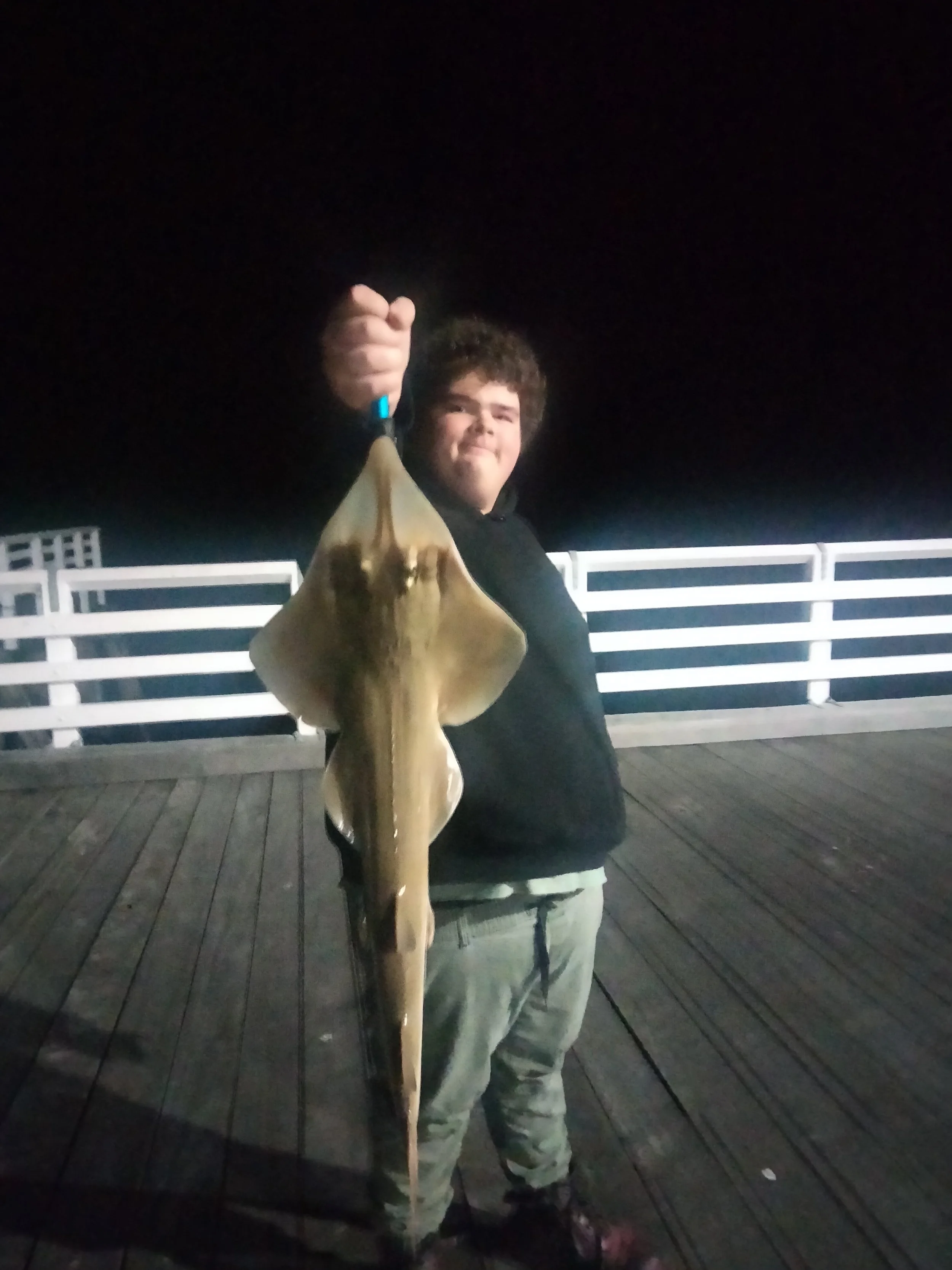 A person with curly hair and light-colored pants holding a large, shiny fish on a wooden pier at night.