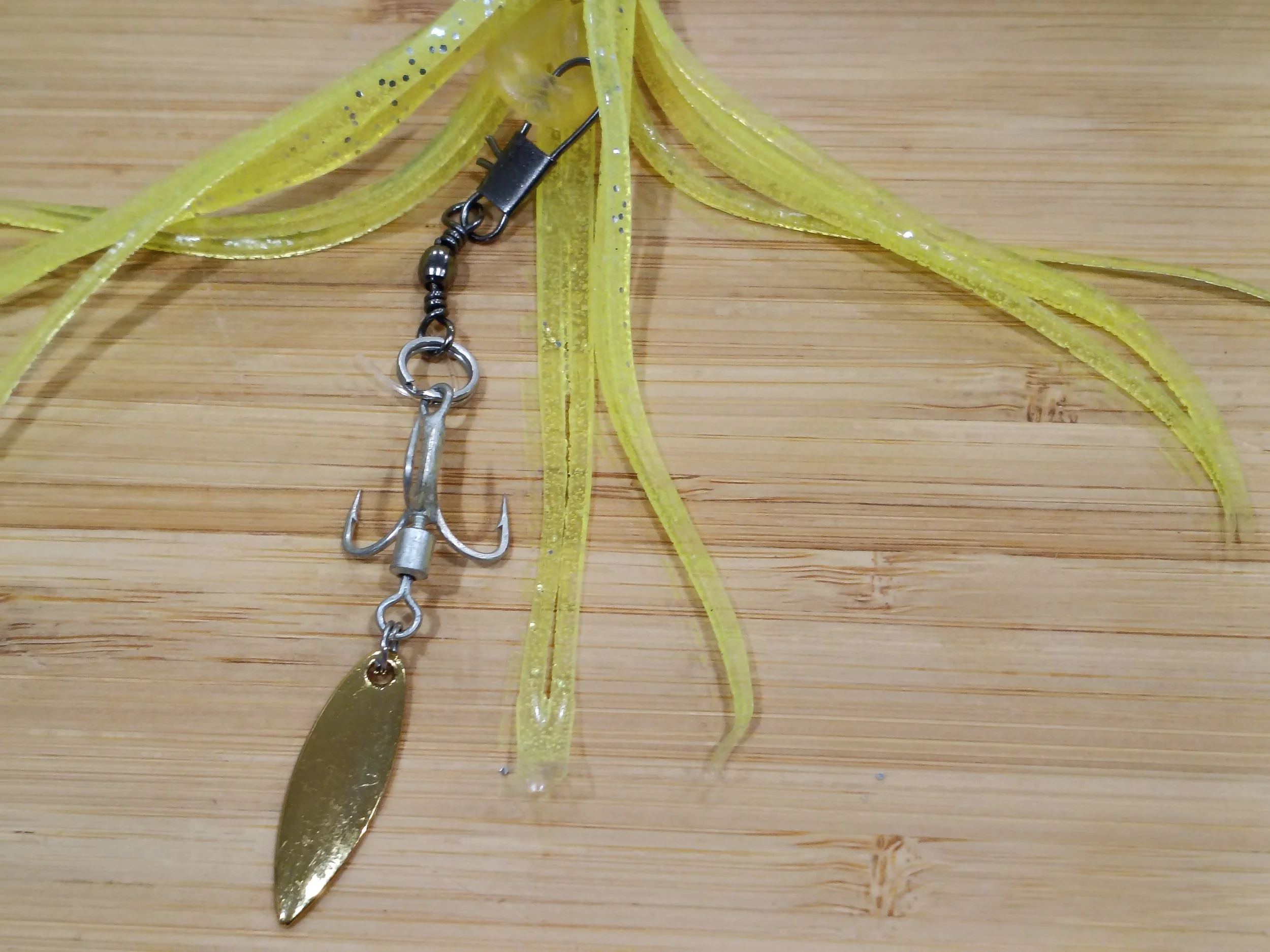 Close-up of several yellow soft plastic fishing lures with silver glitter, hooks, and a fishing sinker on a wooden surface.