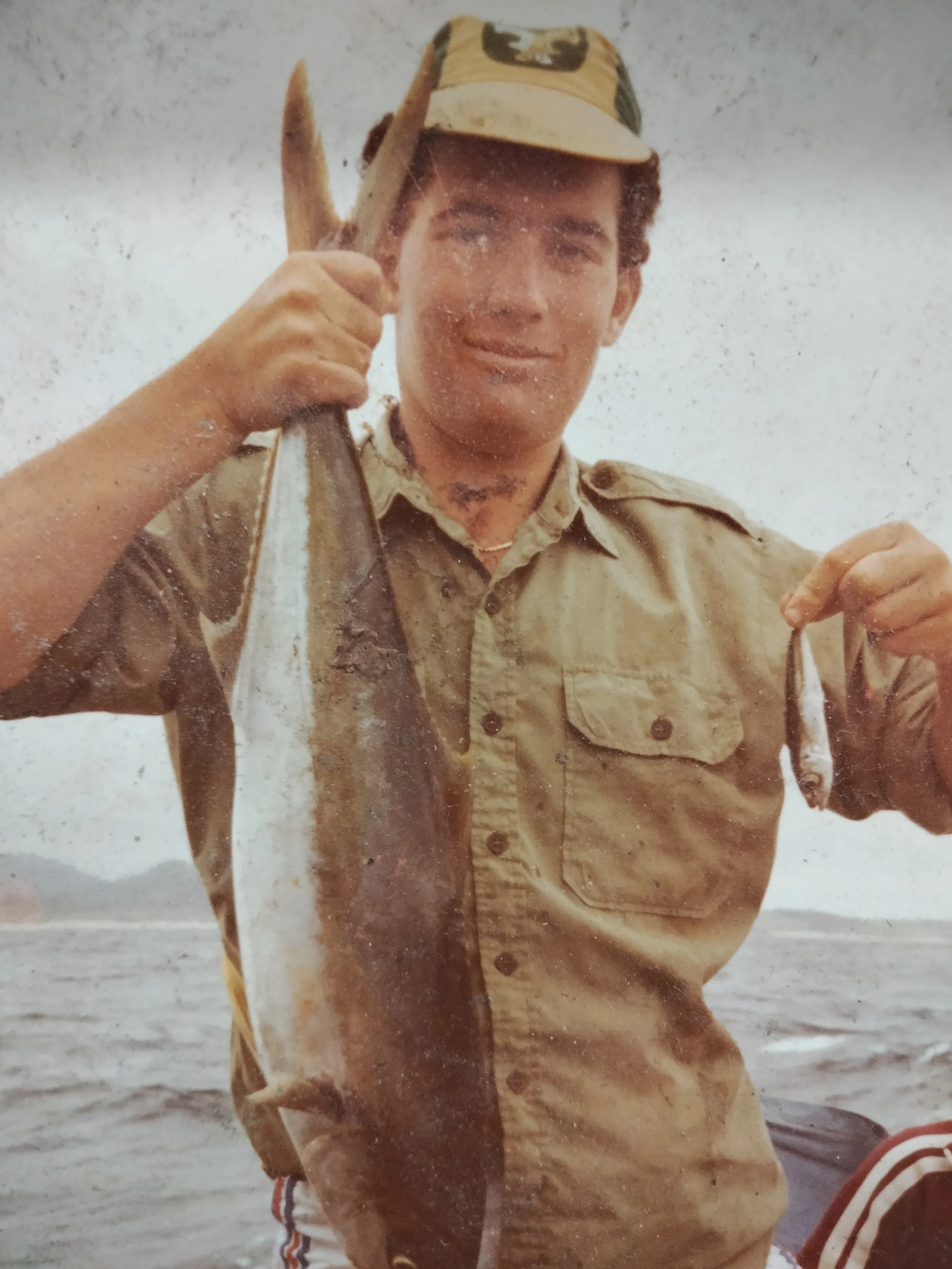 A young man in beige military uniform holding a fish in his right hand at a beach or shoreline.