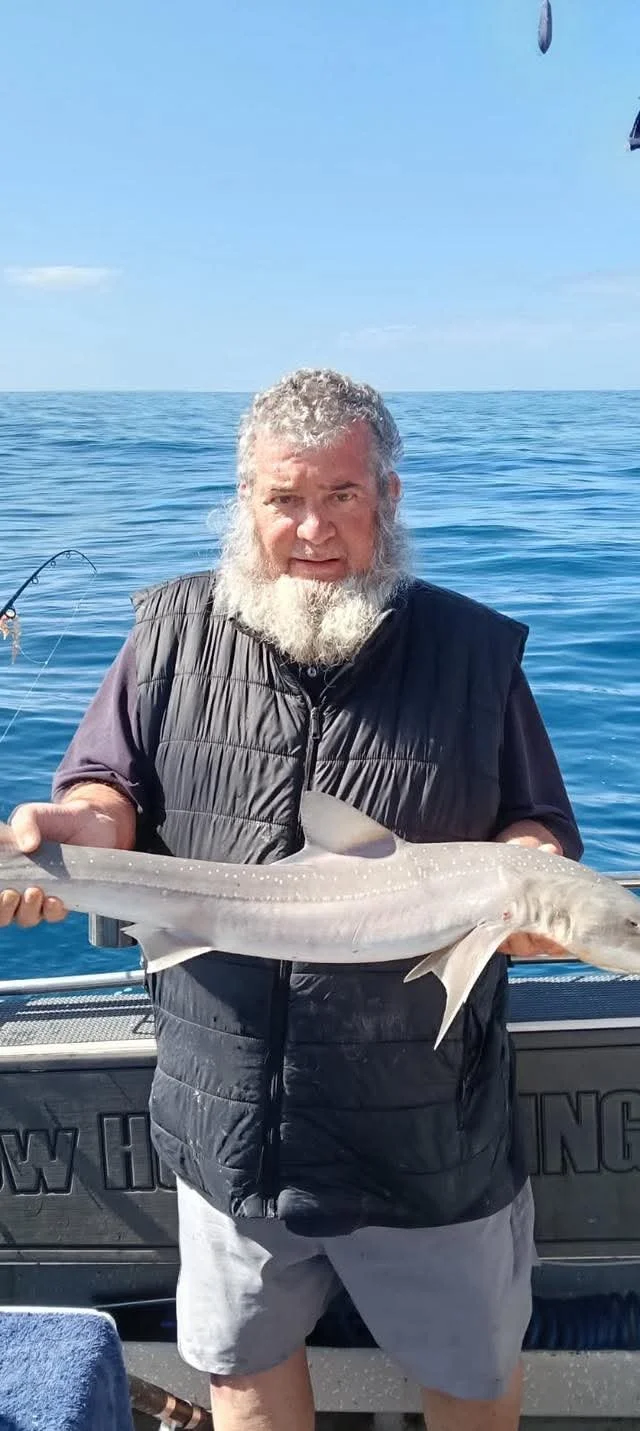 A man with a gray beard, wearing a black vest and gray shorts, holding a large fish on a boat at sea.
