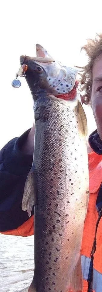 Person holding a large spotted fish with its mouth open, showing a fishing lure hanging from the fish's mouth, outdoors near a body of water.