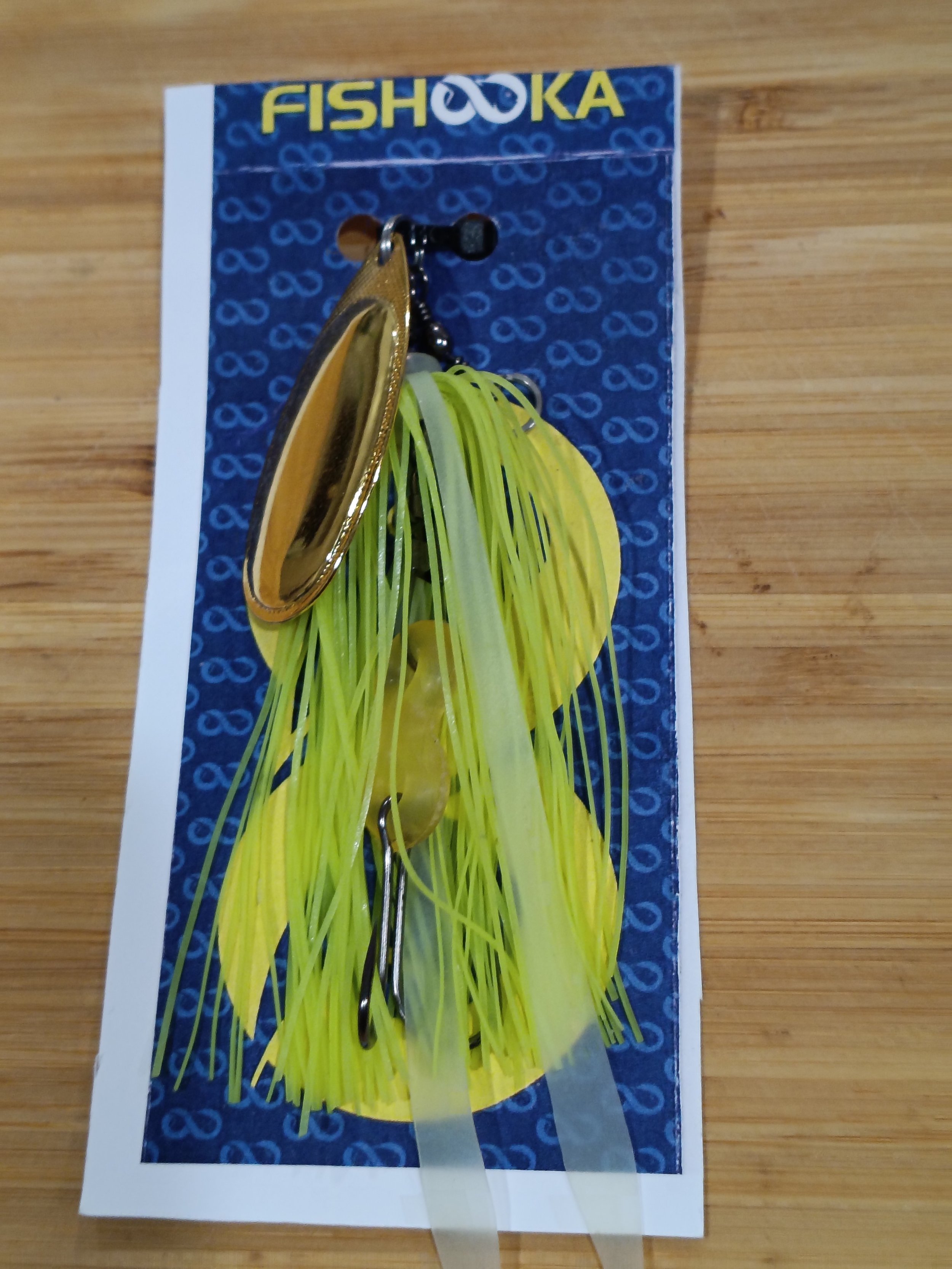 A fishing lure with yellow skirt and a metallic gold body attached to a card with 'FISHOOKA' branding, on a wooden surface.