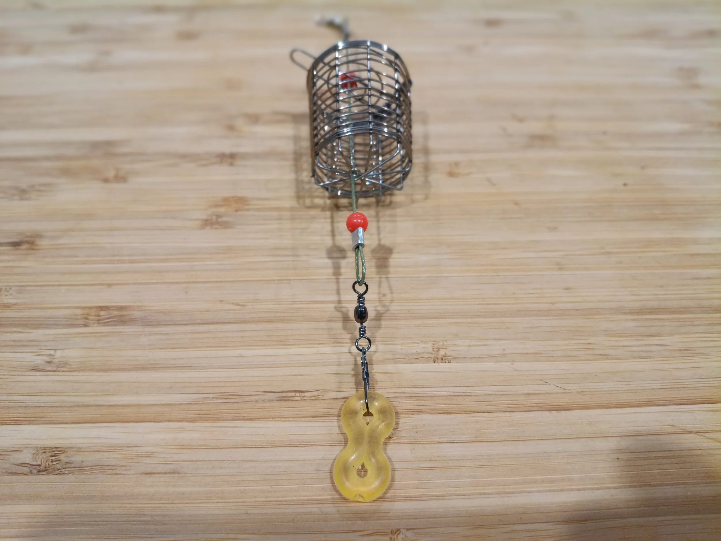 A metal cylindrical cage hanging from a string with a red bead and a yellow doughnut-shaped object attached to it, placed on a wooden surface.