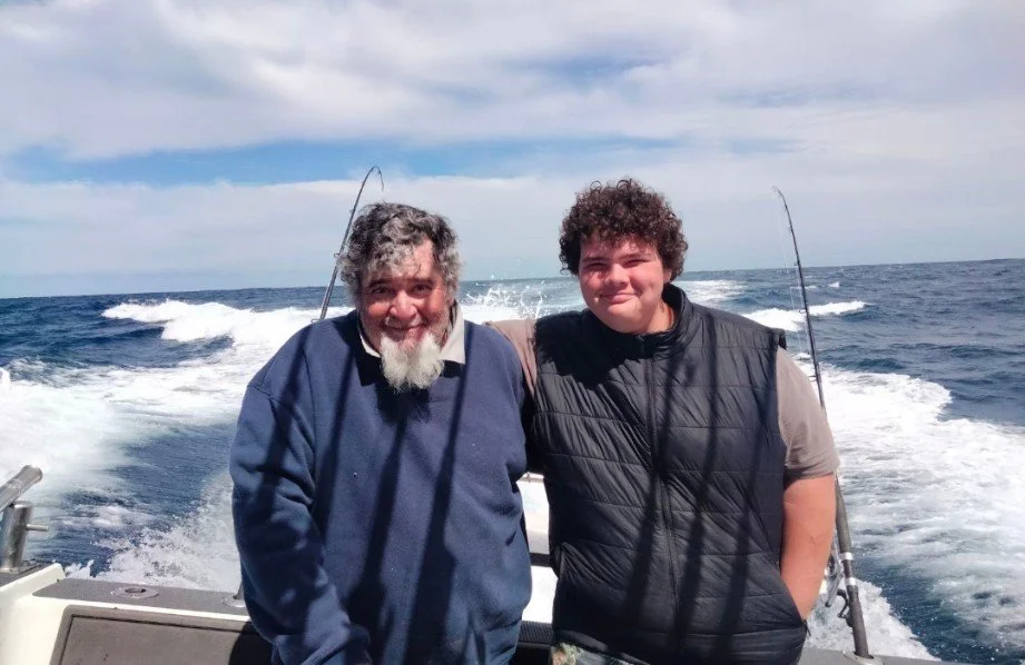Two smiling men on a boat, with the ocean and wake in the background, fishing rods behind them.