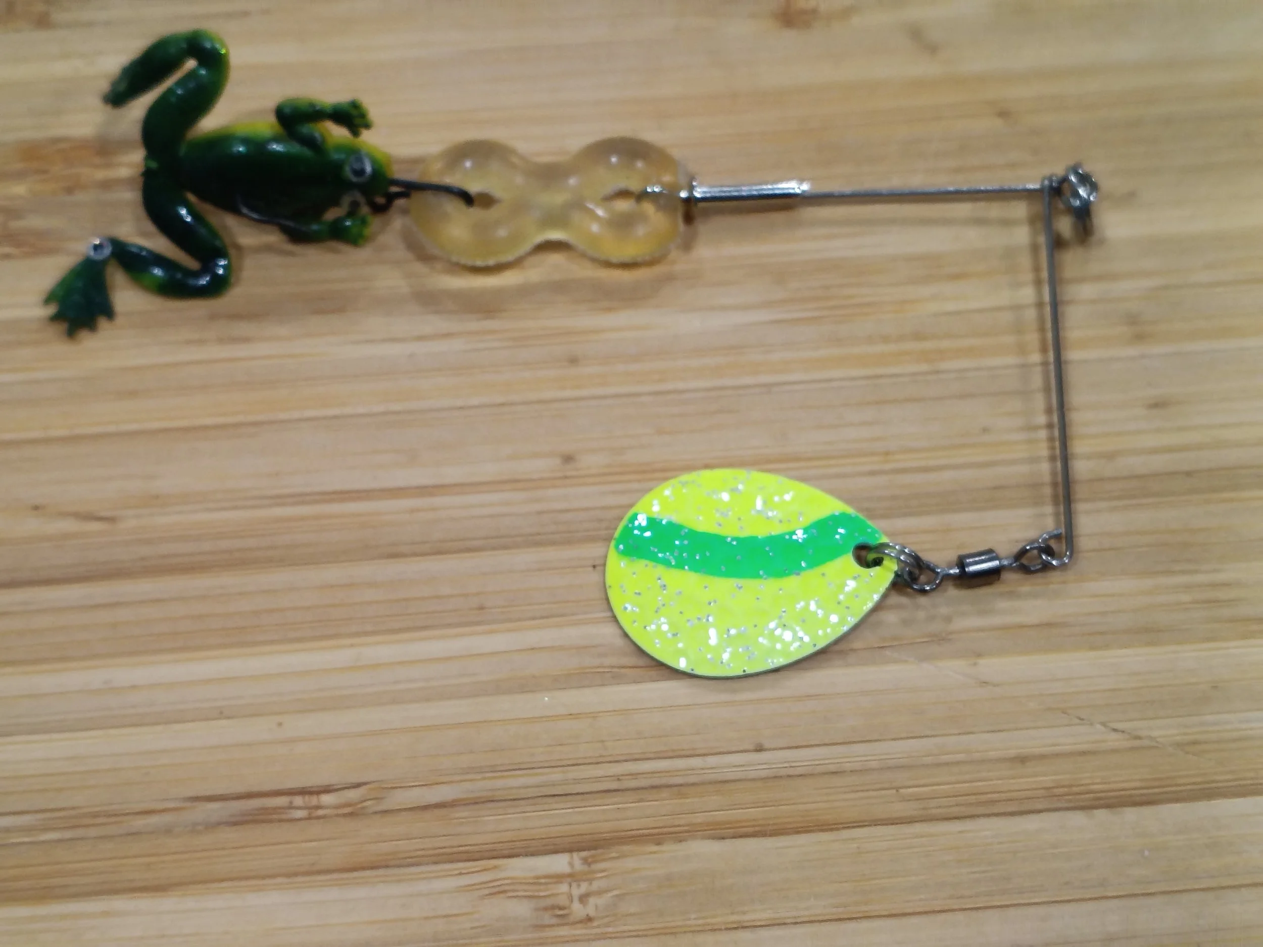 A fishing lure designed to resemble a frog with green and black coloring, attached to a metal hook and chain, lying on a wooden surface.