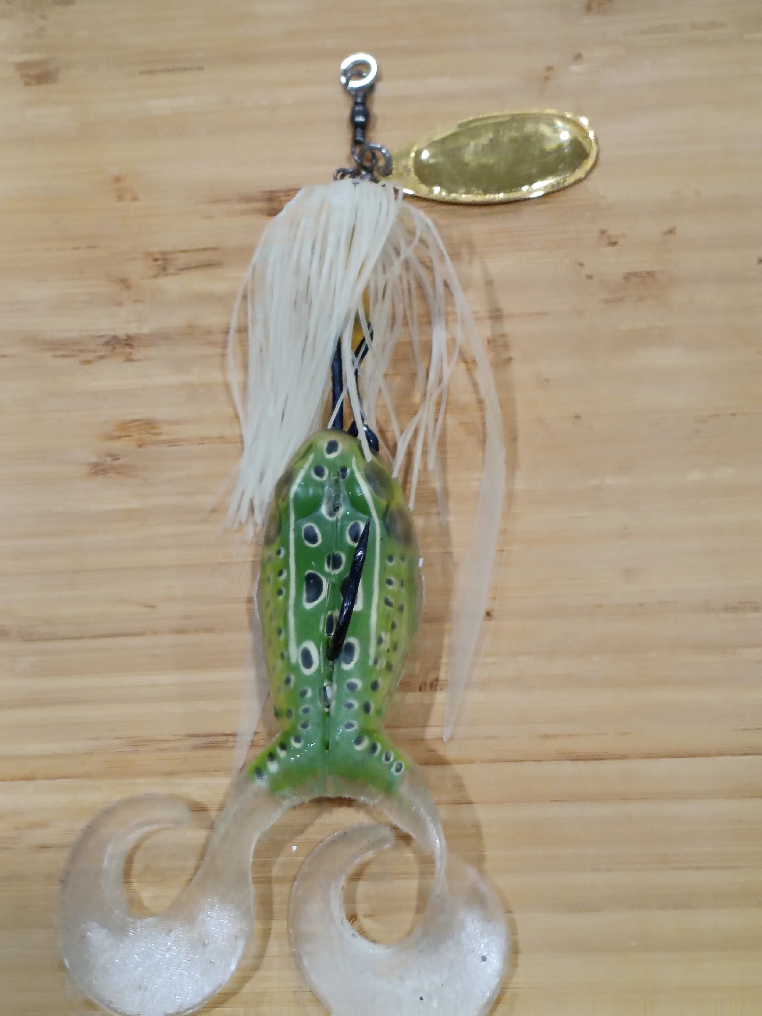 A fishing lure with a frog design and feather details, attached to a chain and a metallic oval-shaped tag, resting on a wooden surface.