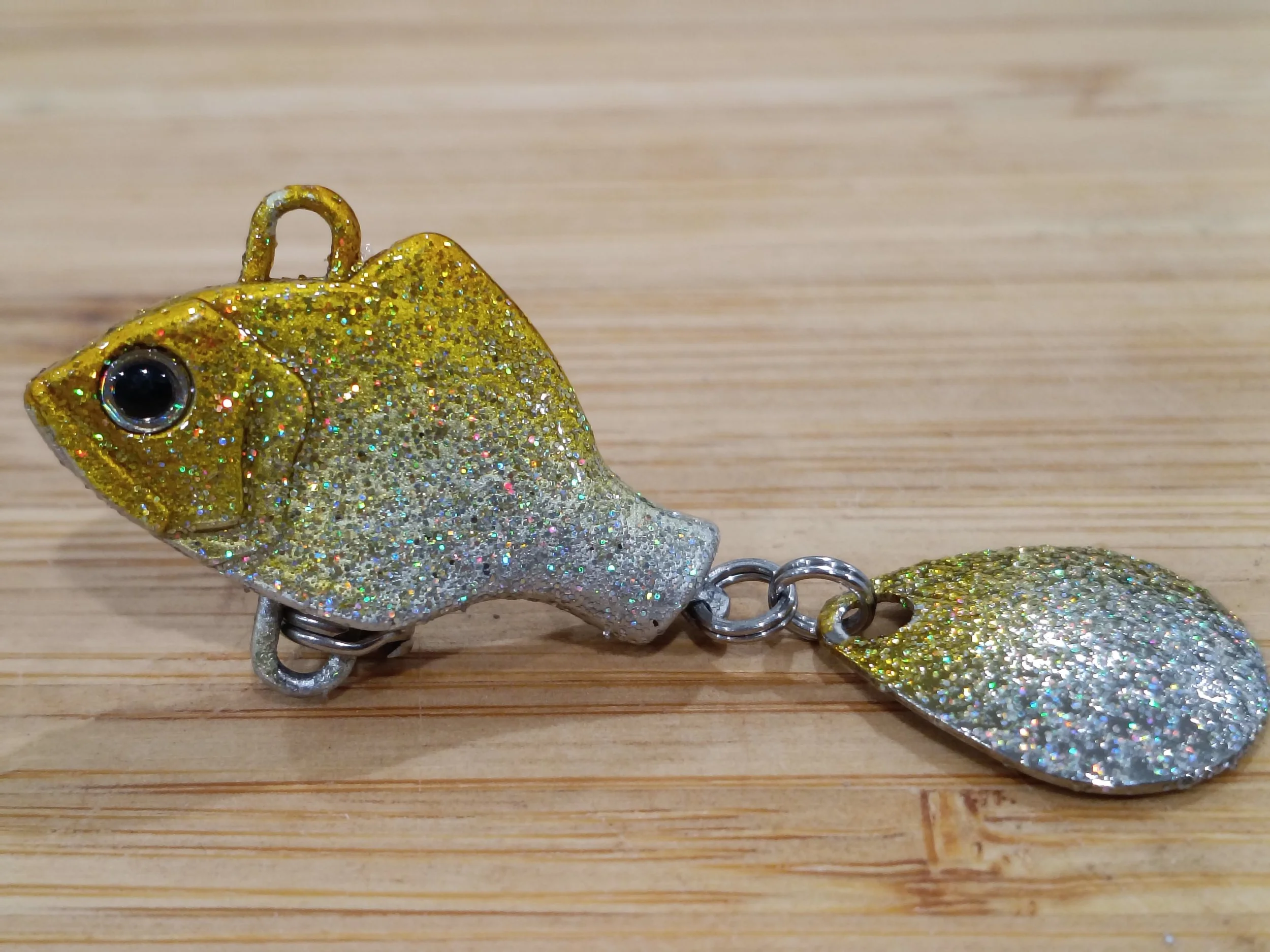 Close-up of a glittery, gold and silver fish-shaped keychain with a large black eye, lying on a wooden surface.