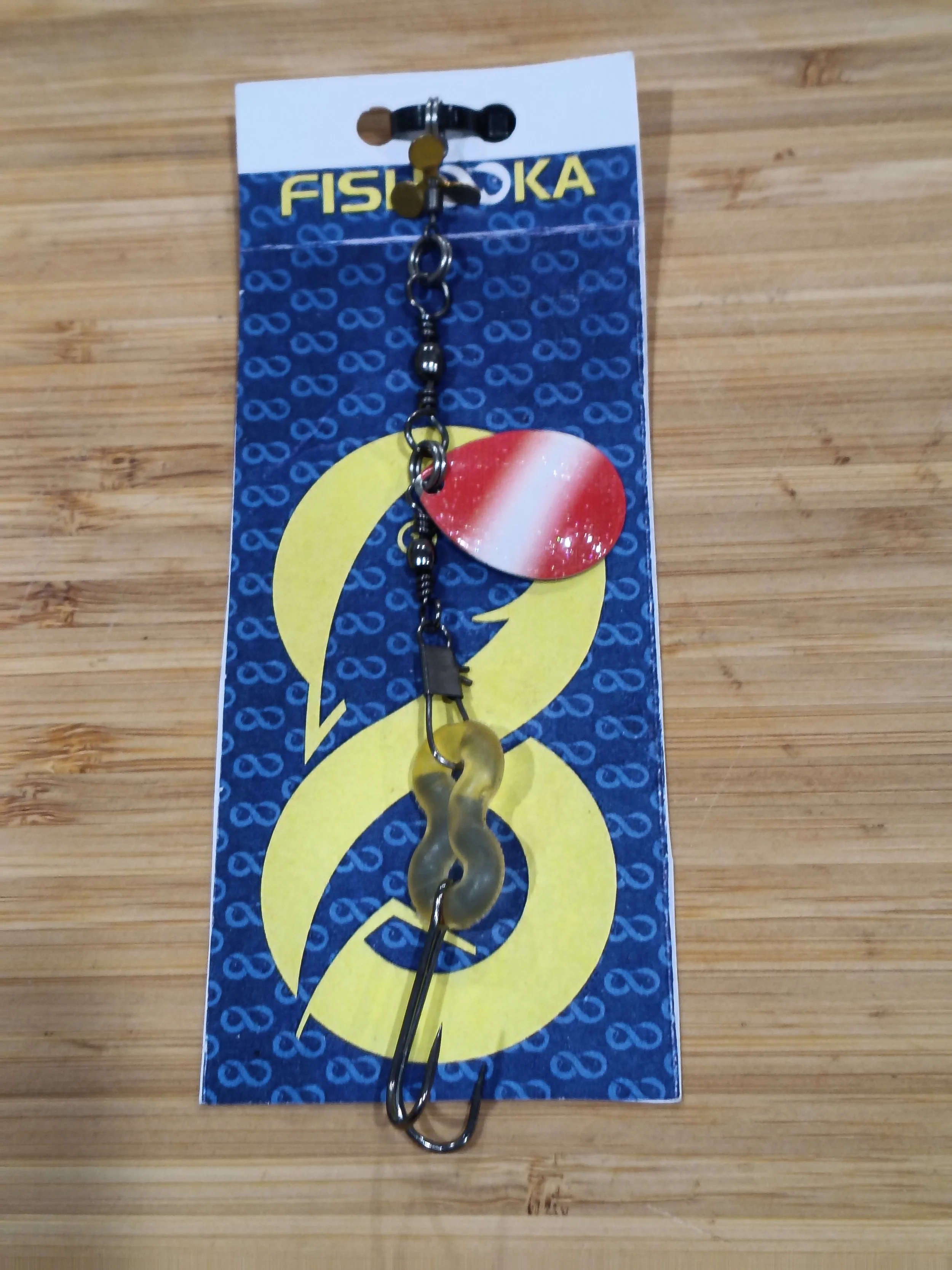 Fishing lure with a red and white spoon attached, displayed on a fishing tackle packaging with a blue and yellow design, on a wooden surface.