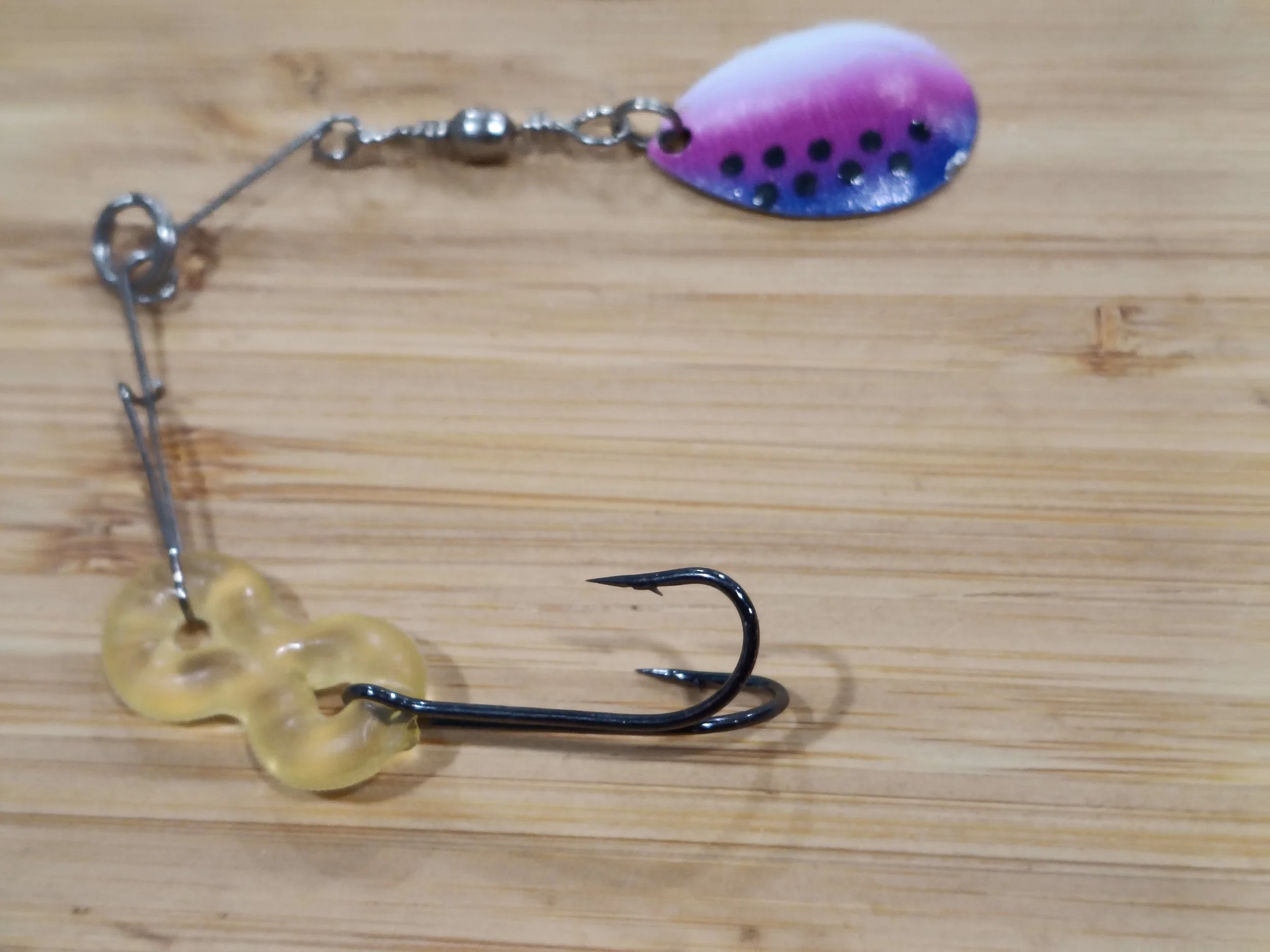 Close-up of a fishing lure with a colorful fish scale pattern, featuring a pink, purple, and blue gradient and black dots, attached to a metal hook on a wooden surface.