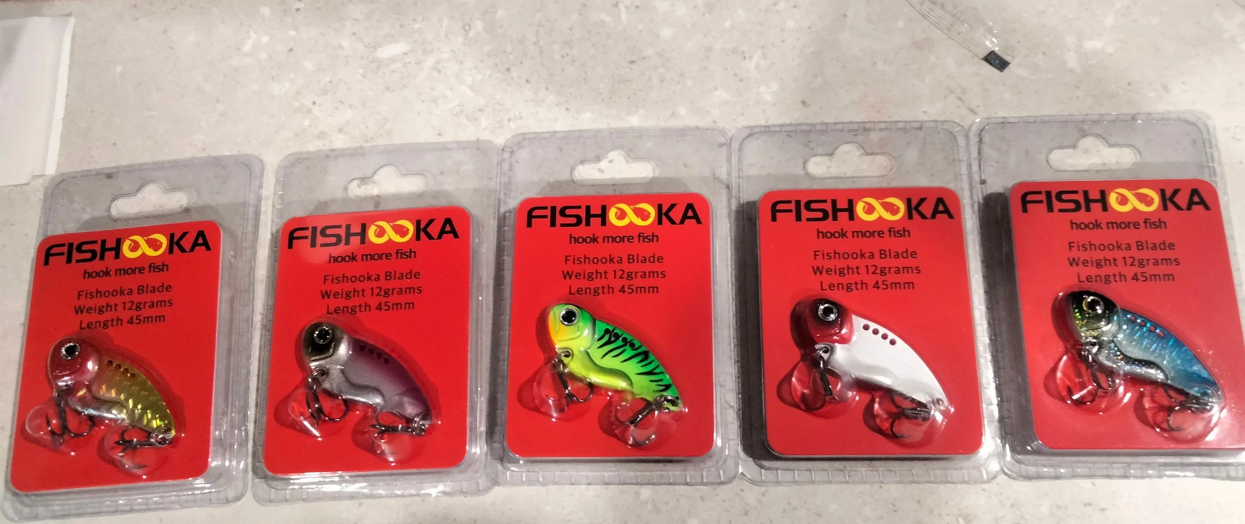 Five fishing lures in plastic packaging, each labeled 'FISHOKA' with the slogan 'hook more fish', with specifications: weight 12 grams and length 45mm.