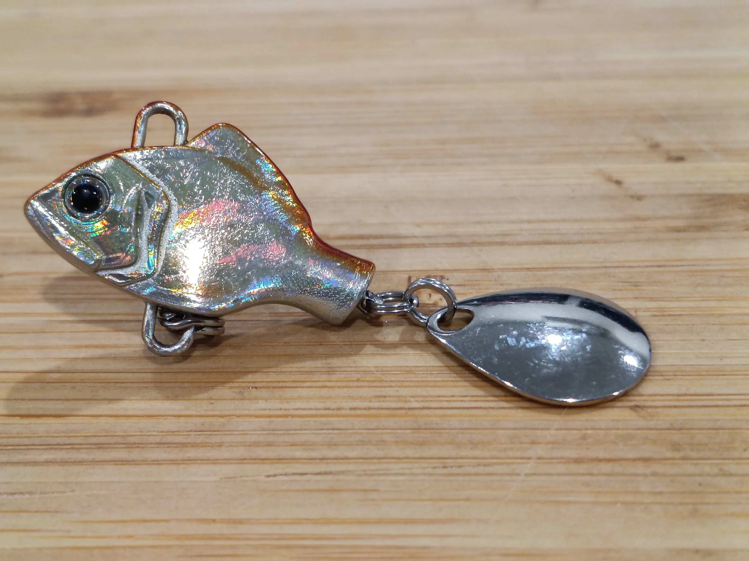 A small, iridescent fish-shaped keychain with a black eye, attached to a metal chain and spoon, lying on a wooden surface.